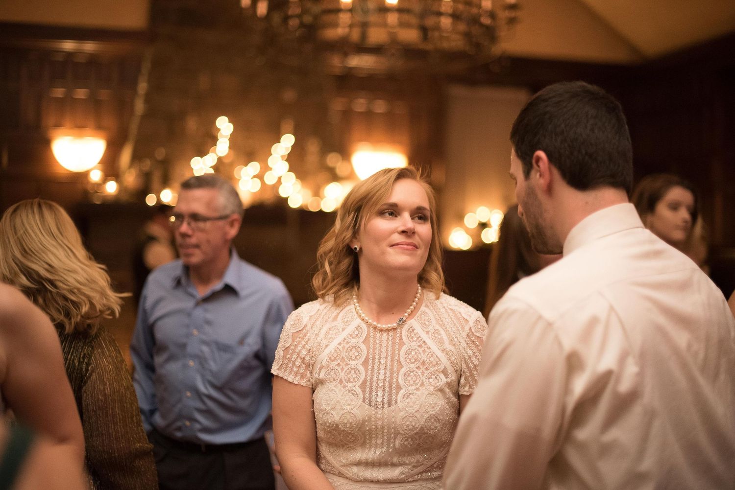 Woman in lace dress smiling at a man during an elegant indoor evening reception with warm chandelier lighting.