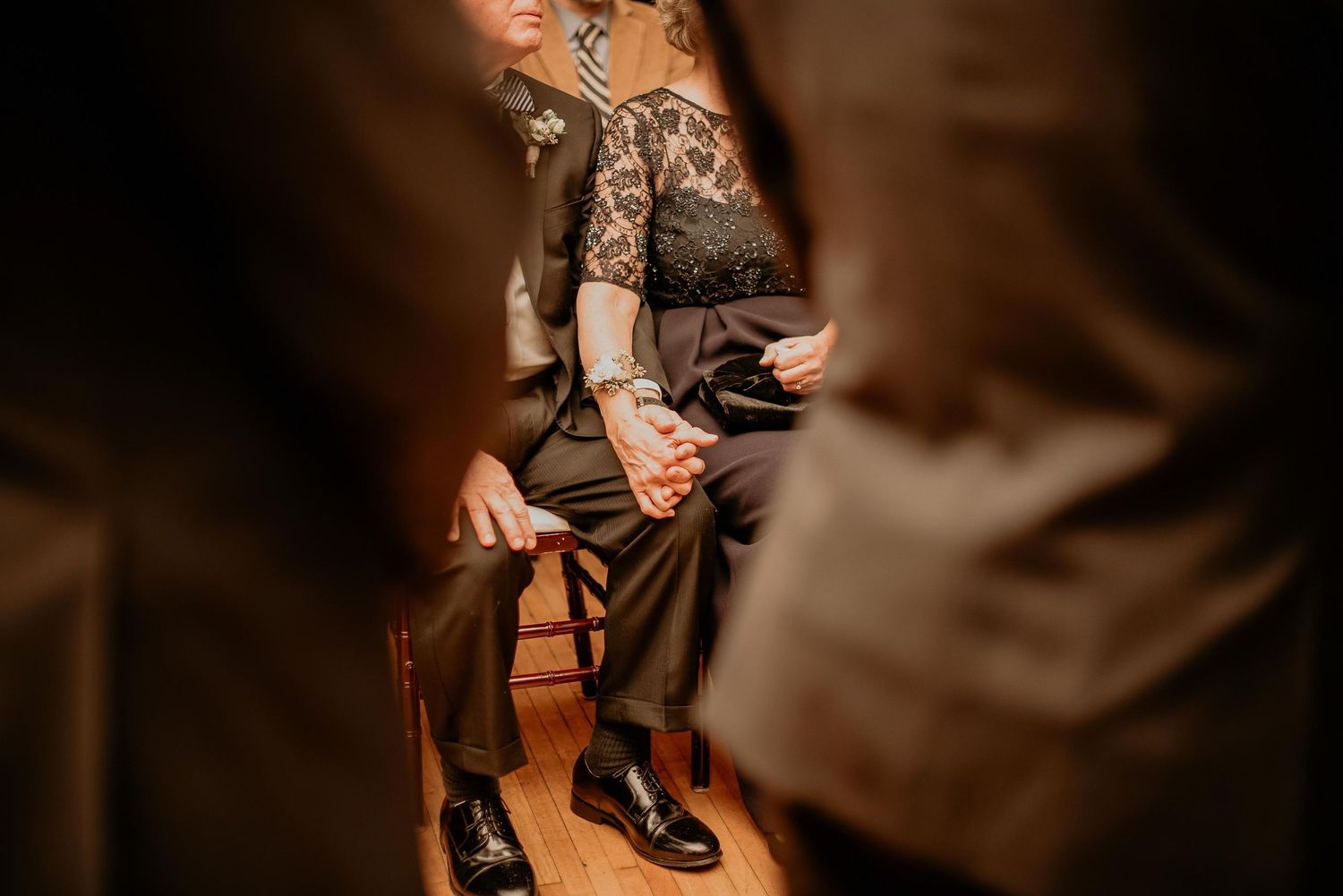 Person seated in chair wearing lace top and dress pants with tattooed arms, photographed through blurred foreground figures.