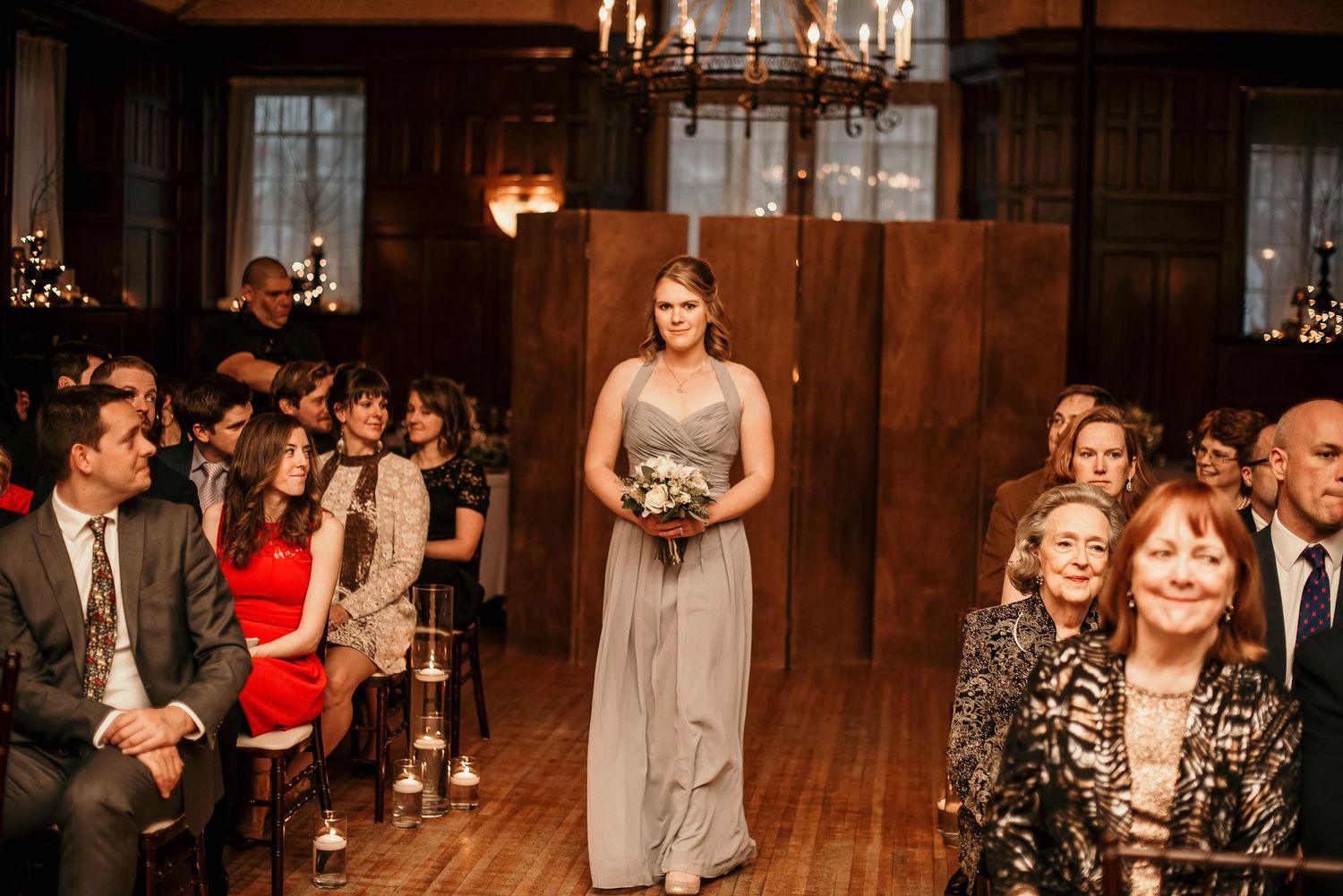 Bridesmaid in gray gown walks down the aisle at an elegant wood-paneled indoor wedding ceremony.