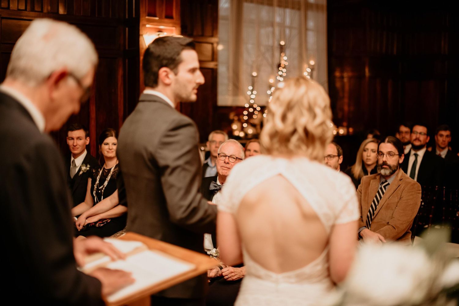 Bride and groom exchange vows at an elegant indoor wedding ceremony with guests seated in a warmly lit venue.