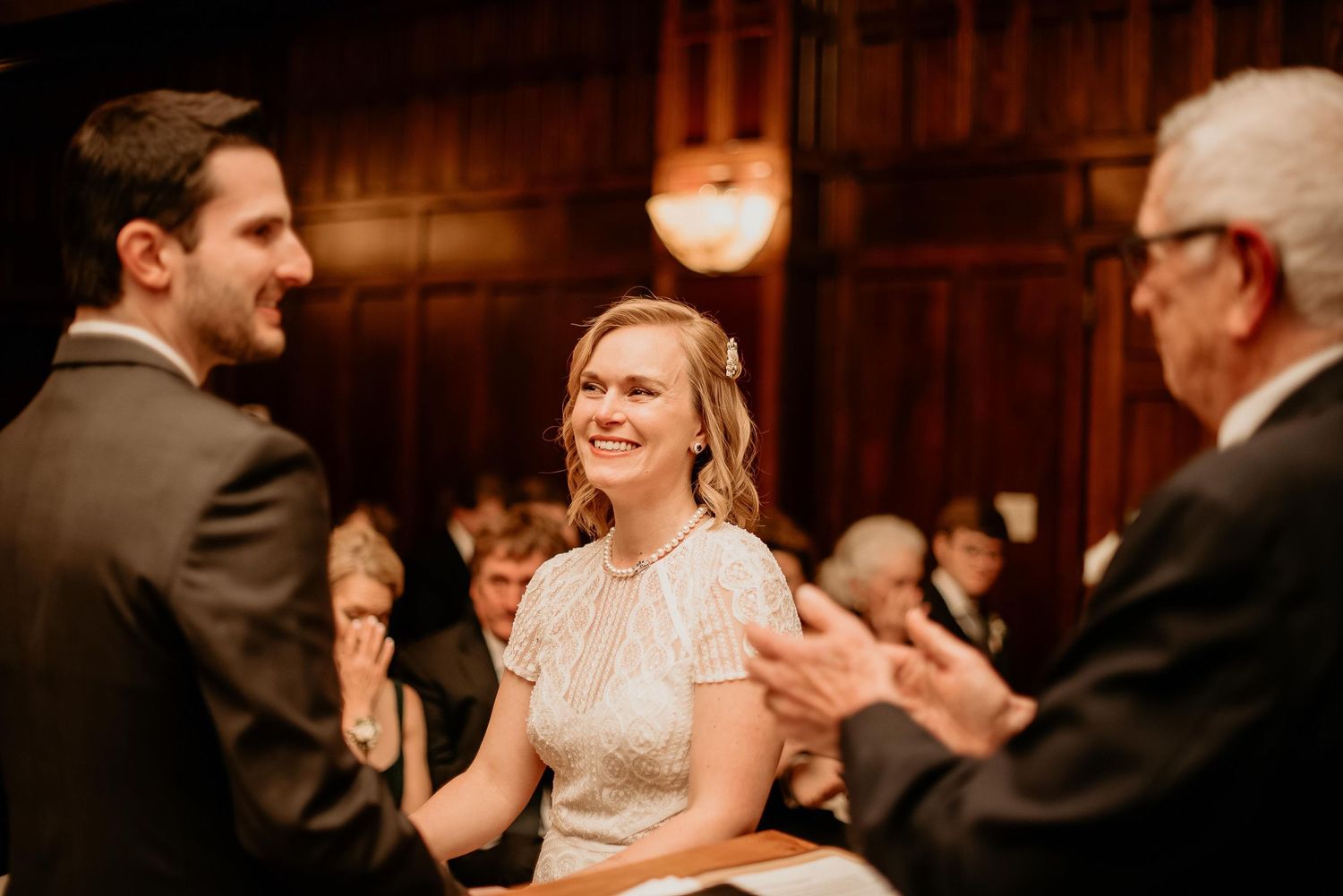 Bride smiling joyfully during wedding ceremony in elegant wood-paneled venue with guests applauding in background.