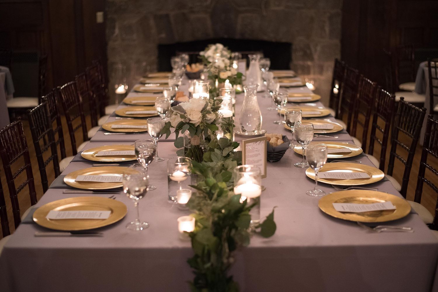 Elegant wedding reception table with gold chargers, white roses, greenery runner, candles, and dark wood chairs by a stone fireplace.