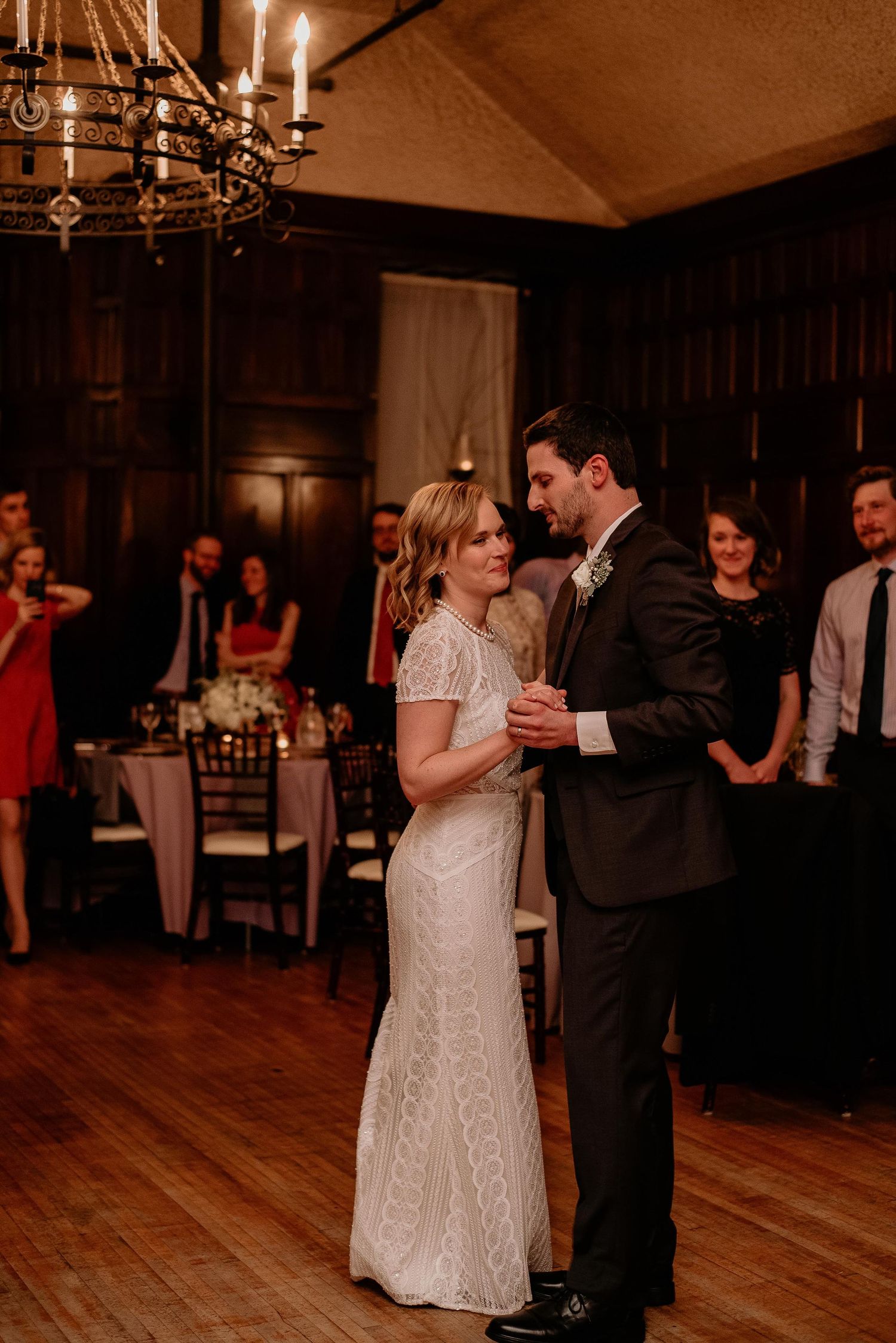 Bride and groom share their first dance in an elegant wood-paneled ballroom with chandelier lighting at their wedding reception.