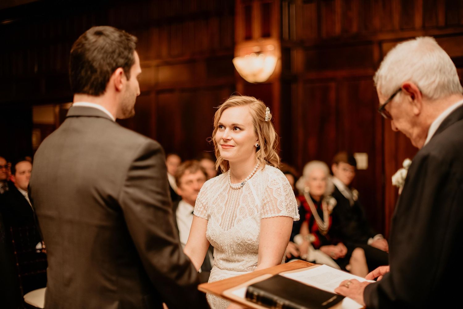 Bride smiling joyfully at groom during intimate wedding ceremony in elegant wood-paneled venue.