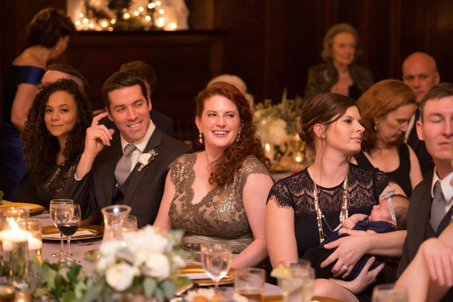 Guests smiling and laughing at an elegant candlelit wedding reception dinner table with floral arrangements.