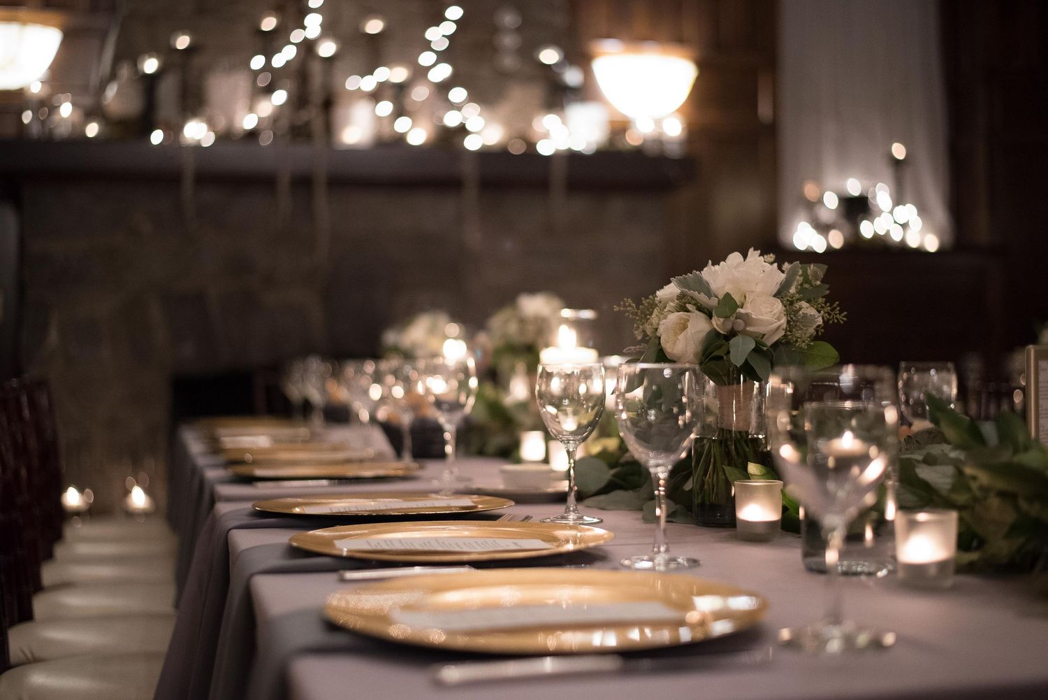 Elegant wedding reception table with gold charger plates, crystal glasses, white floral centerpieces, and soft candlelight ambiance.