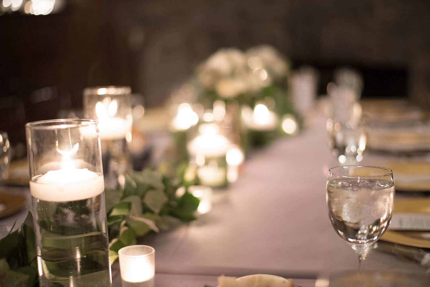 Elegant wedding reception table with floating candles, greenery garland, and glassware in soft candlelight.