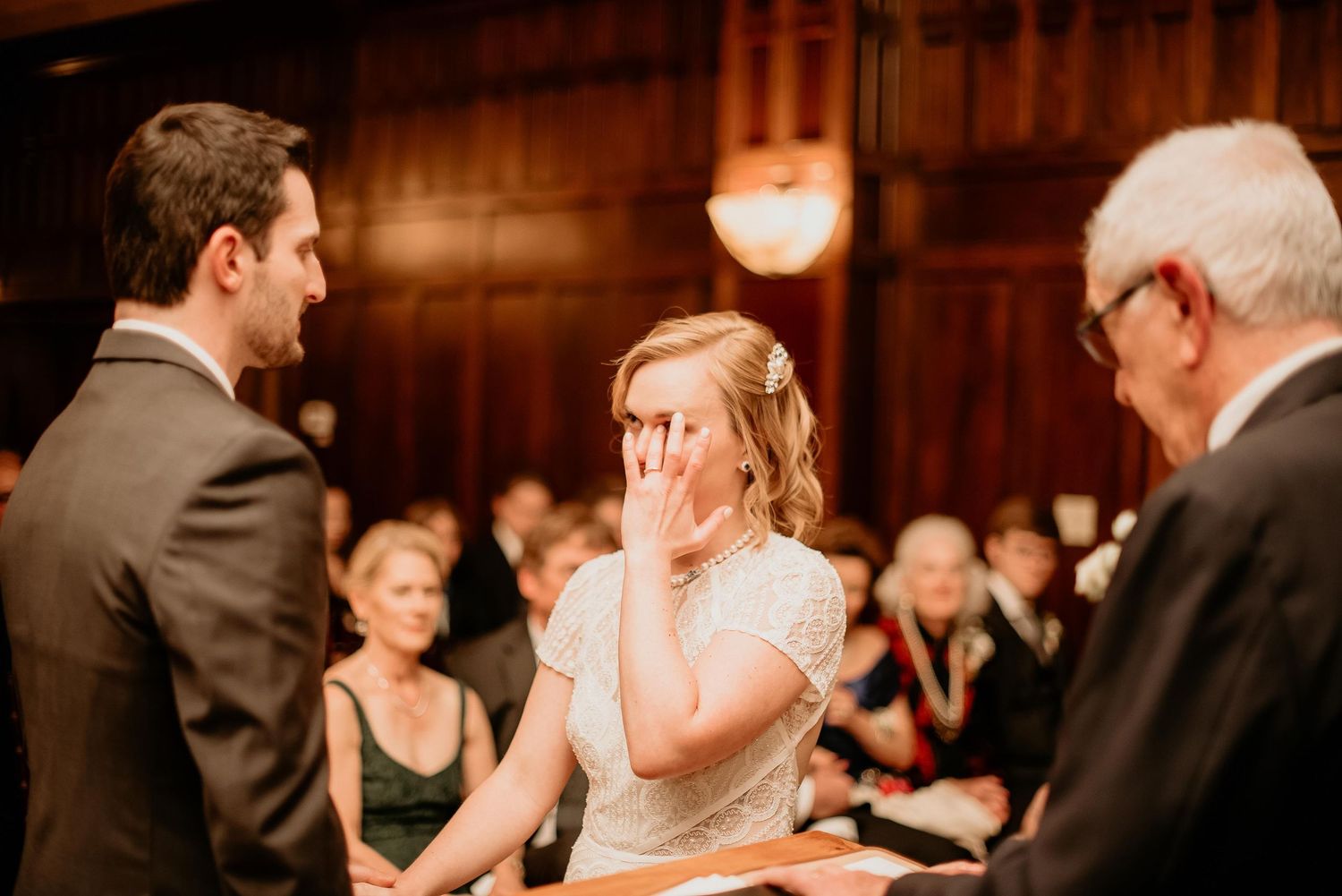 Emotional bride covering her mouth during wedding ceremony while groom and officiant stand nearby in elegant venue.