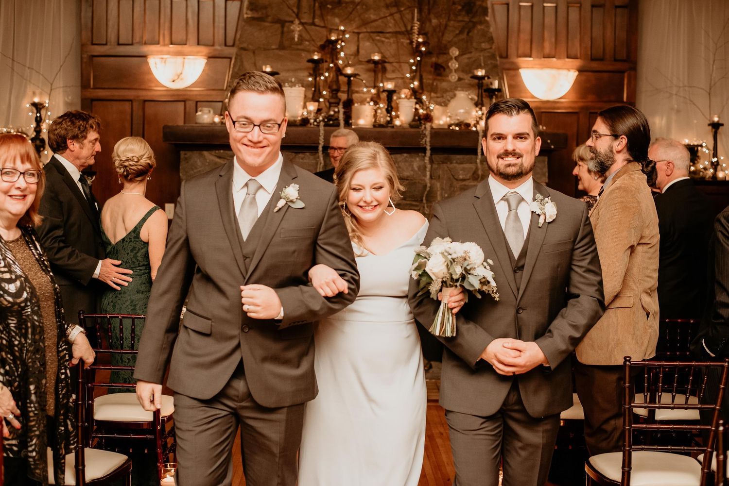 Bride walks down the aisle with two groomsmen in gray suits during an elegant indoor wedding ceremony.