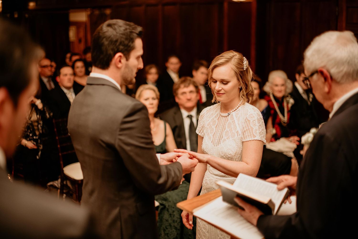 Bride and groom exchanging rings during indoor wedding ceremony with guests seated in background.