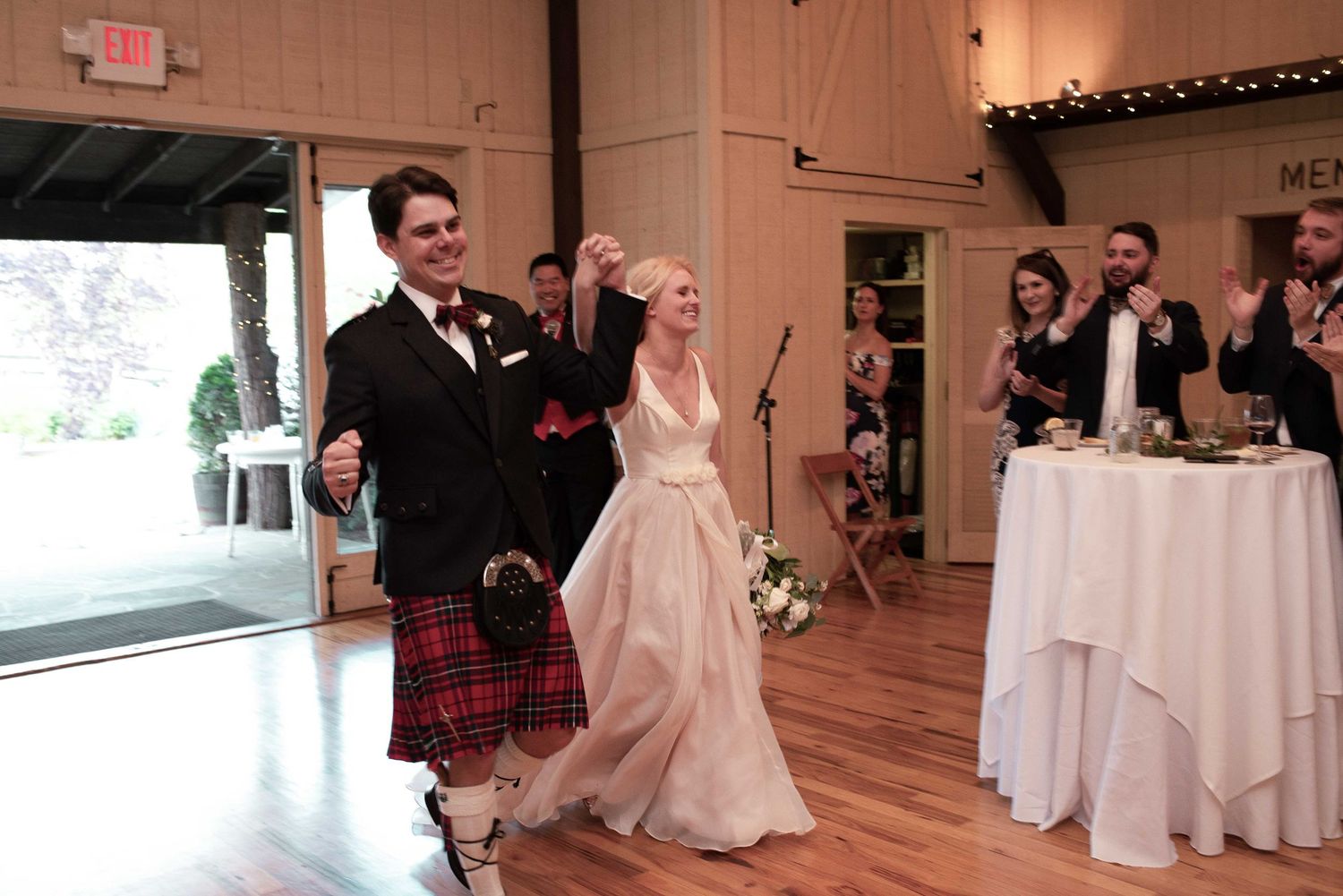 Bride in white gown and groom in red tartan kilt enter wedding reception hall to applause from guests.