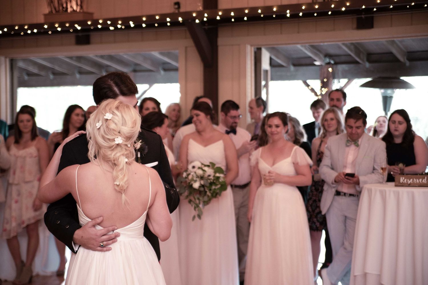 Bride and groom share first dance surrounded by bridesmaids and guests at elegant wedding reception.
