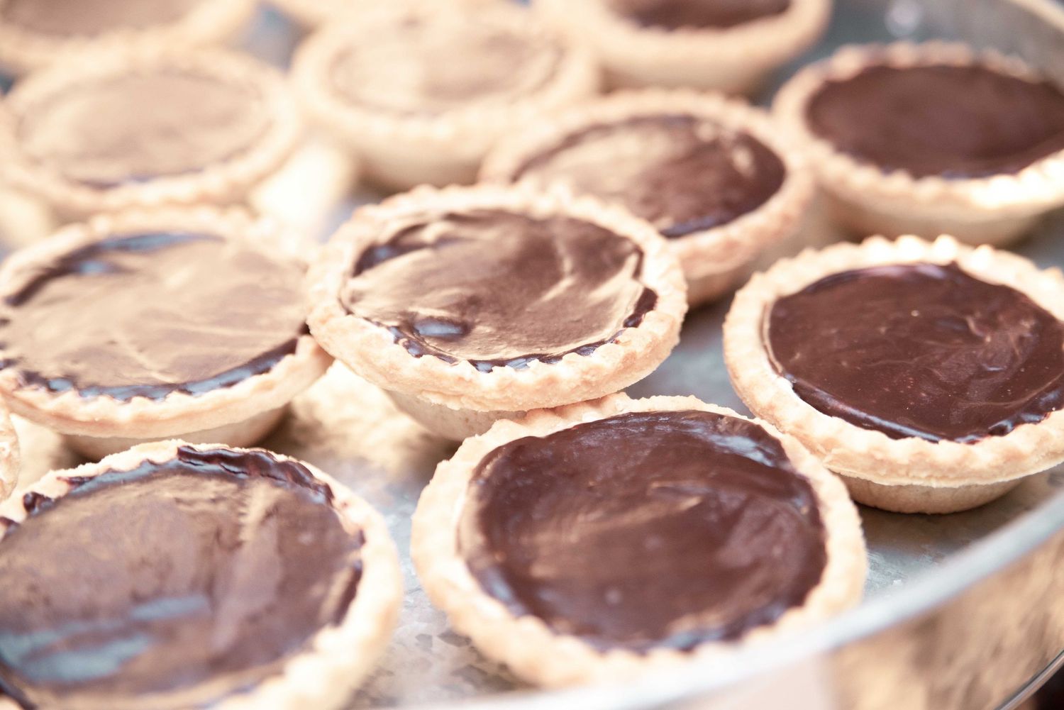 Freshly made chocolate tart shells with glossy ganache filling arranged on a silver baking tray.