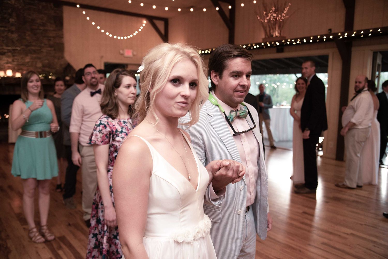 Bride in white dress and groom in gray suit walk the dance floor at a rustic wedding reception venue.