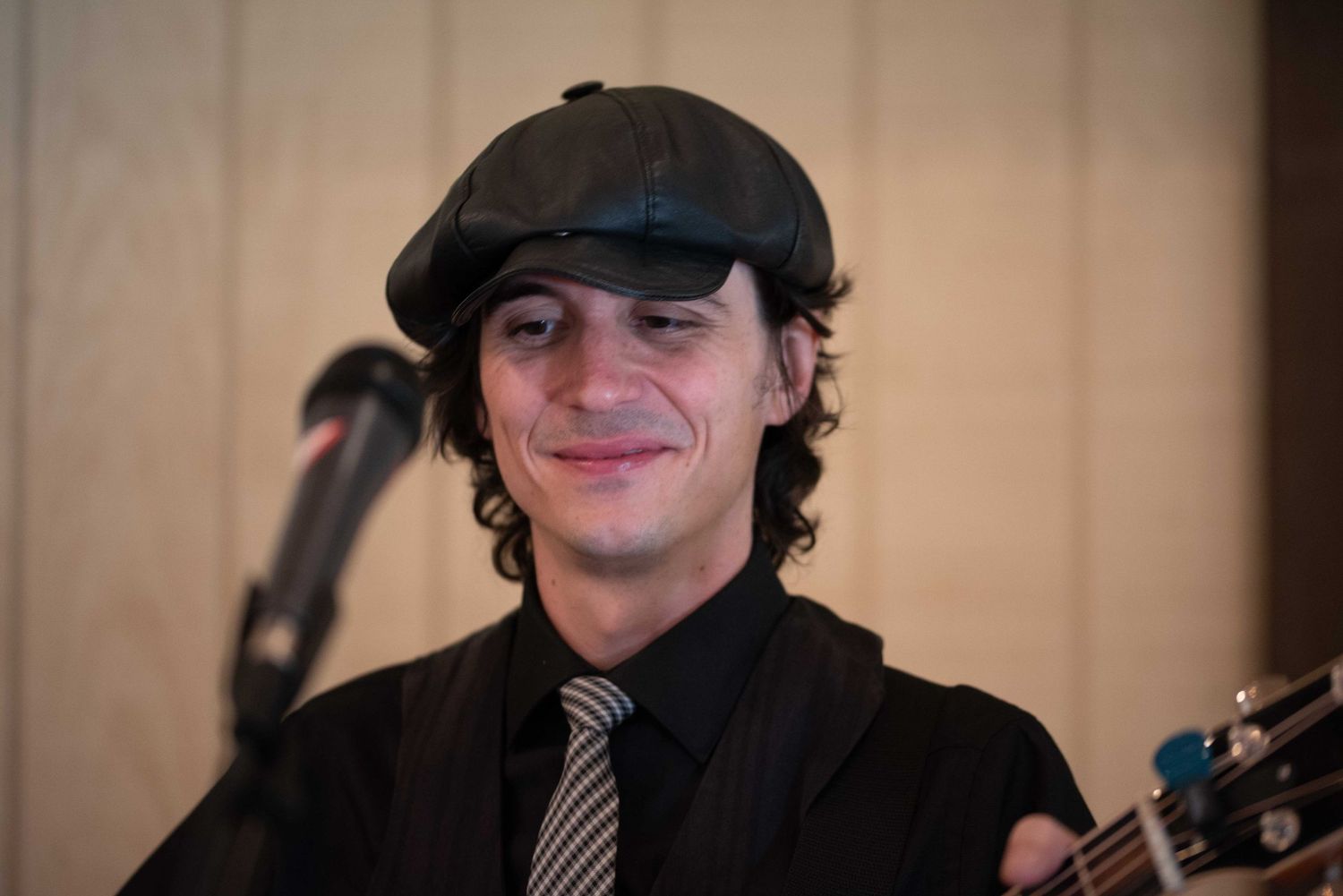 Young man smiling, wearing a flat cap and tie while playing guitar at a live event.