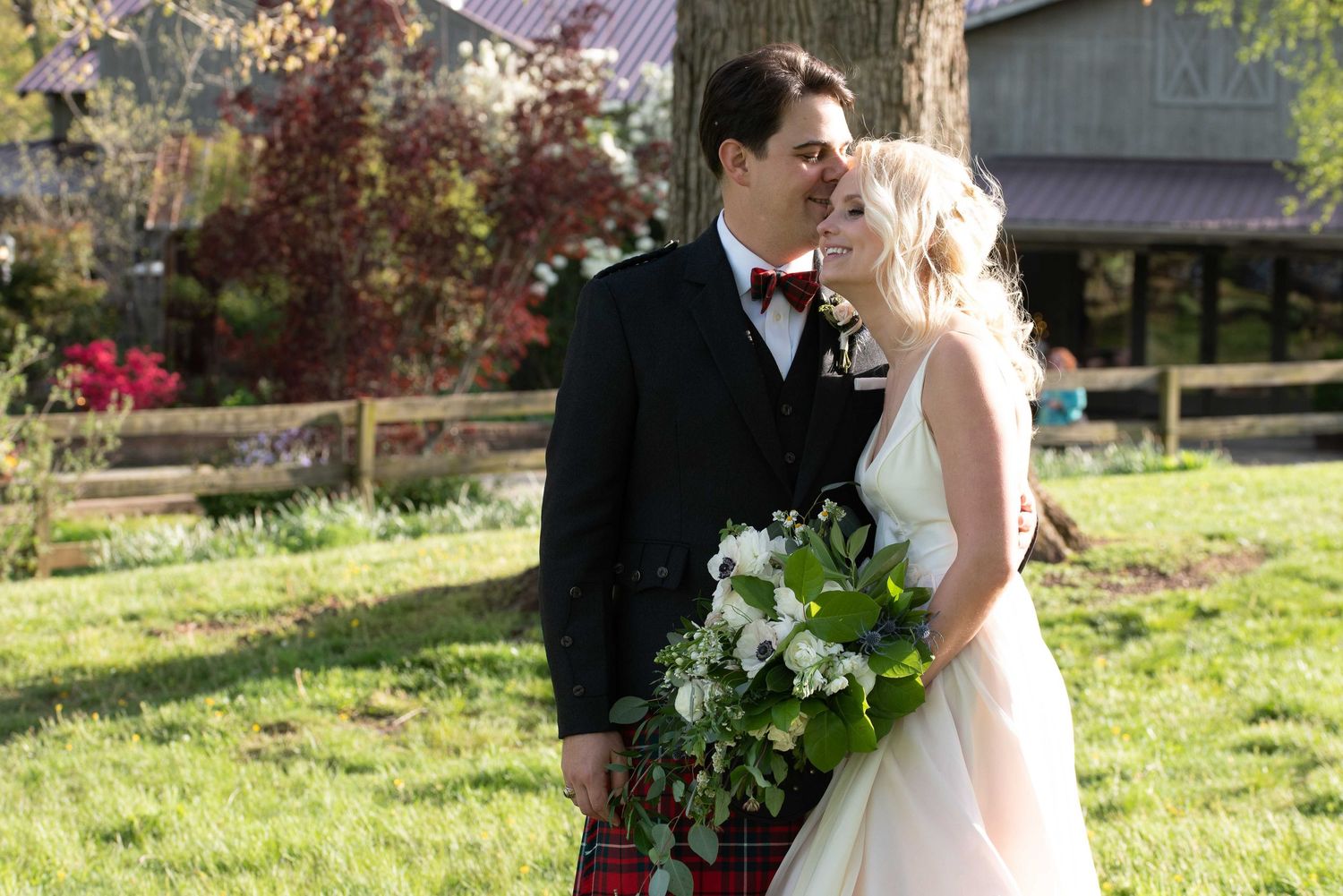 Bride and groom share a romantic kiss outdoors, bride holding white floral bouquet, groom wearing kilt and bow tie.