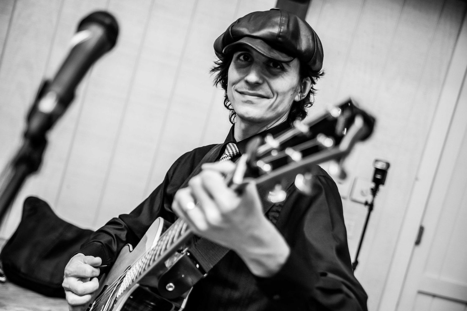 Black and white photo of a musician playing electric guitar, wearing a cap, smiling near a microphone on stage.
