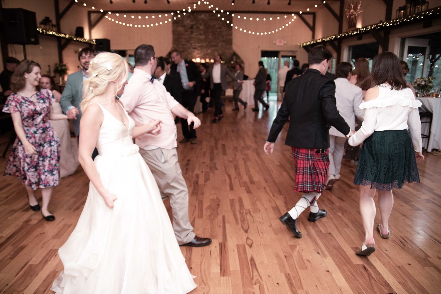 Guests dancing in a circle at a wedding reception on a hardwood floor with string lights overhead.