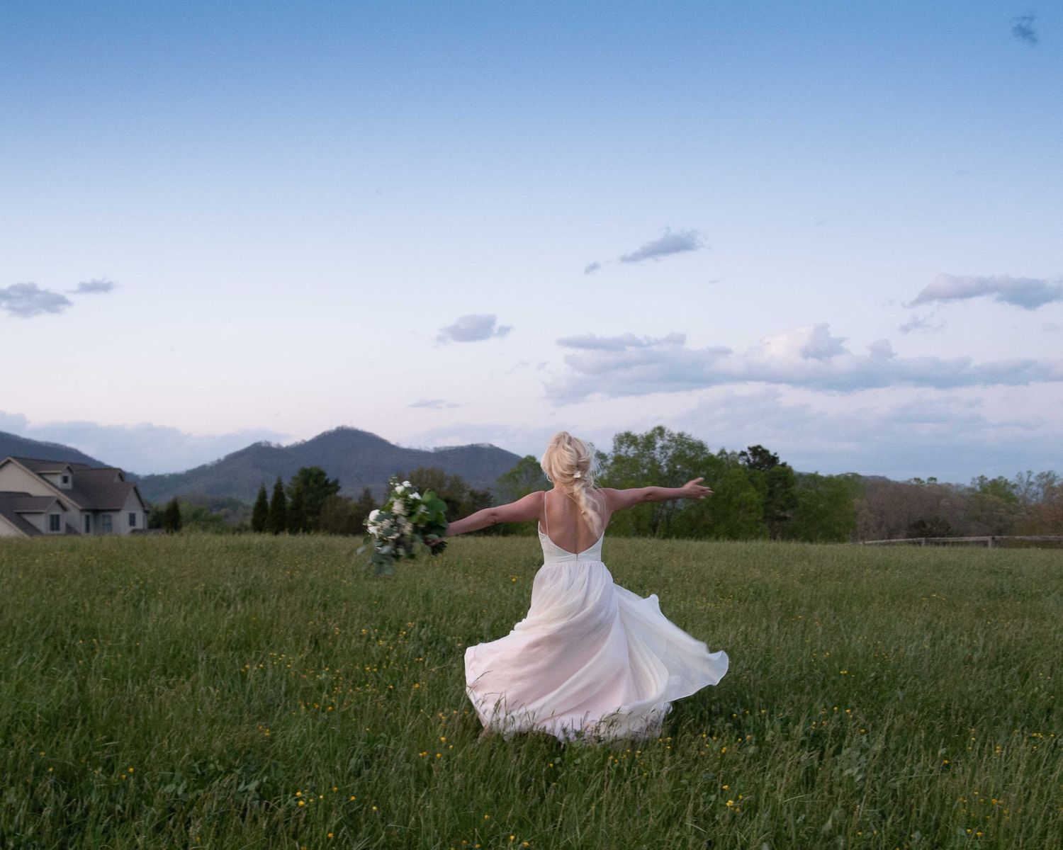 Bride in white gown twirling in green meadow with mountain backdrop at dusk, holding bouquet with arms outstretched.