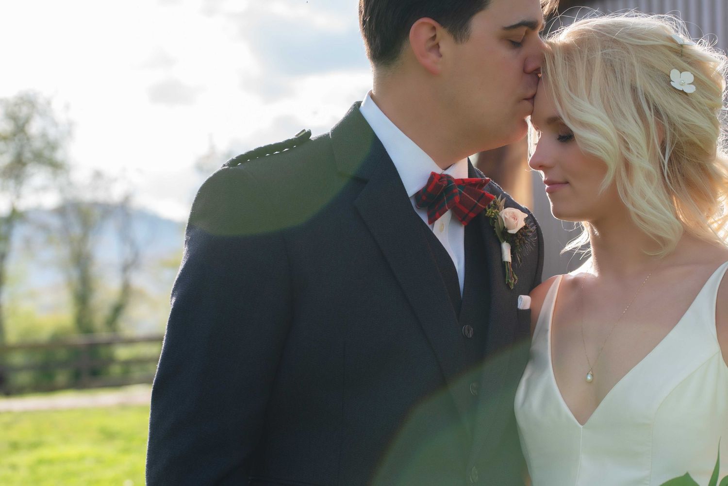 A groom in a navy suit with red bow tie kisses his blonde bride in a white dress outdoors.