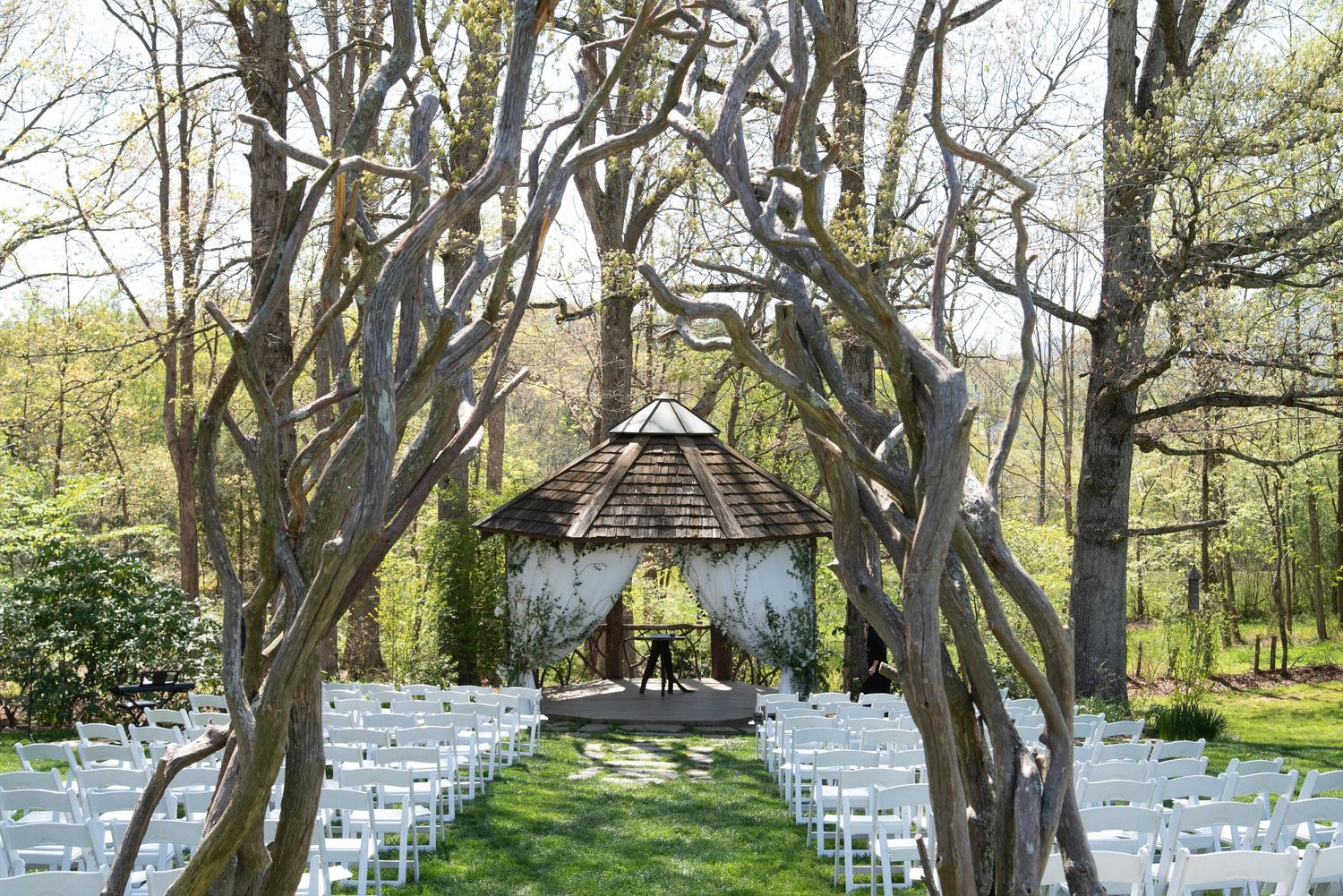 Outdoor wedding ceremony setup with white chairs and rustic gazebo among bare trees.