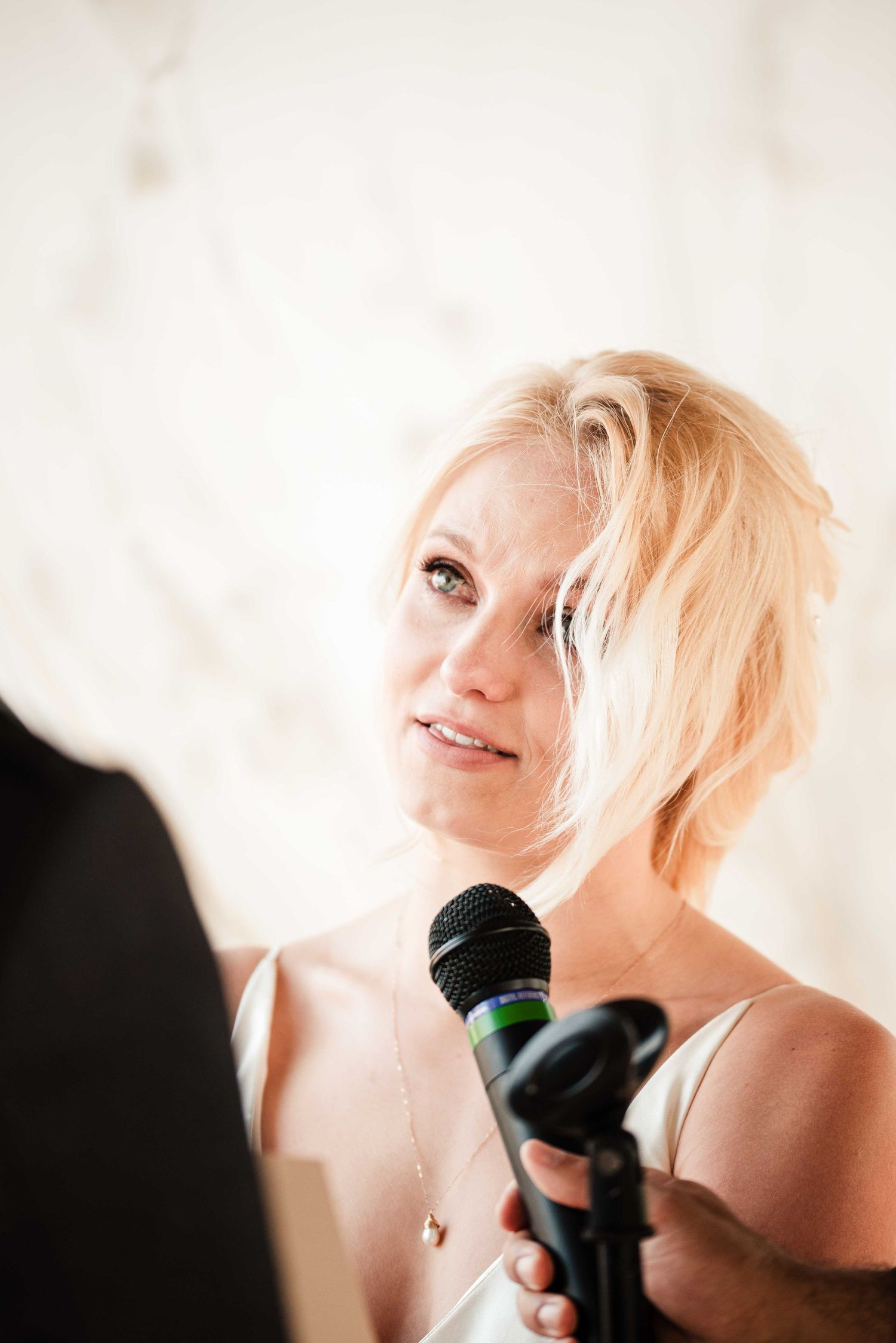 Emotional blonde bride holding a microphone during wedding ceremony speech with tears in her eyes.