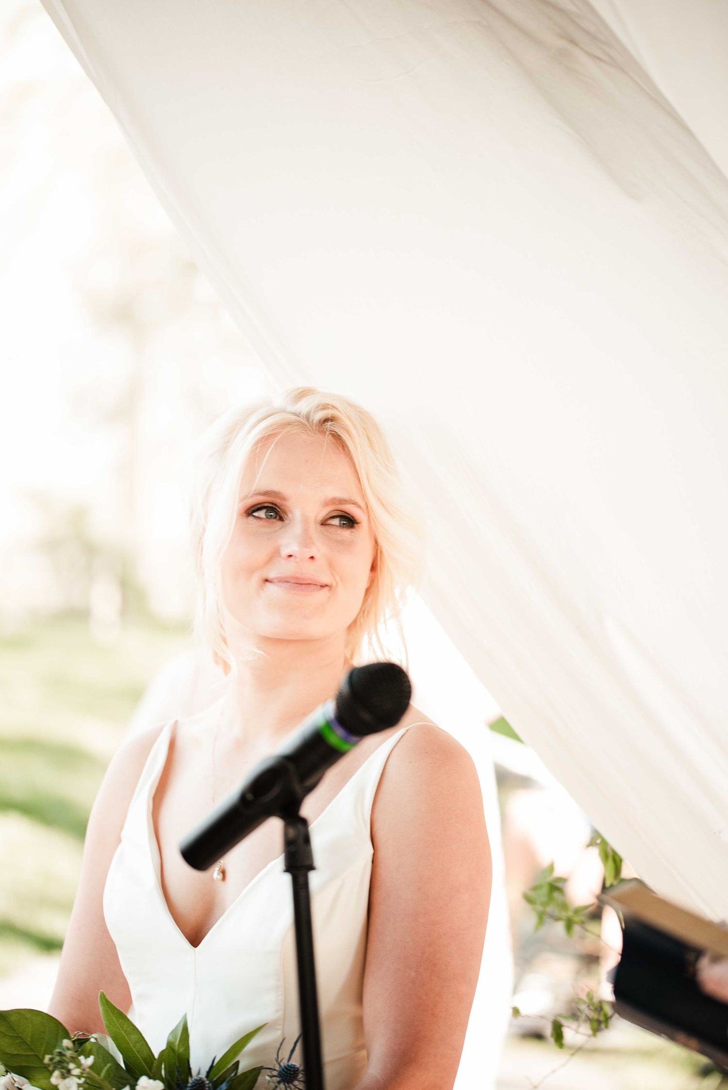 Blonde bride in white gown smiles softly near microphone during outdoor wedding ceremony under white tent.