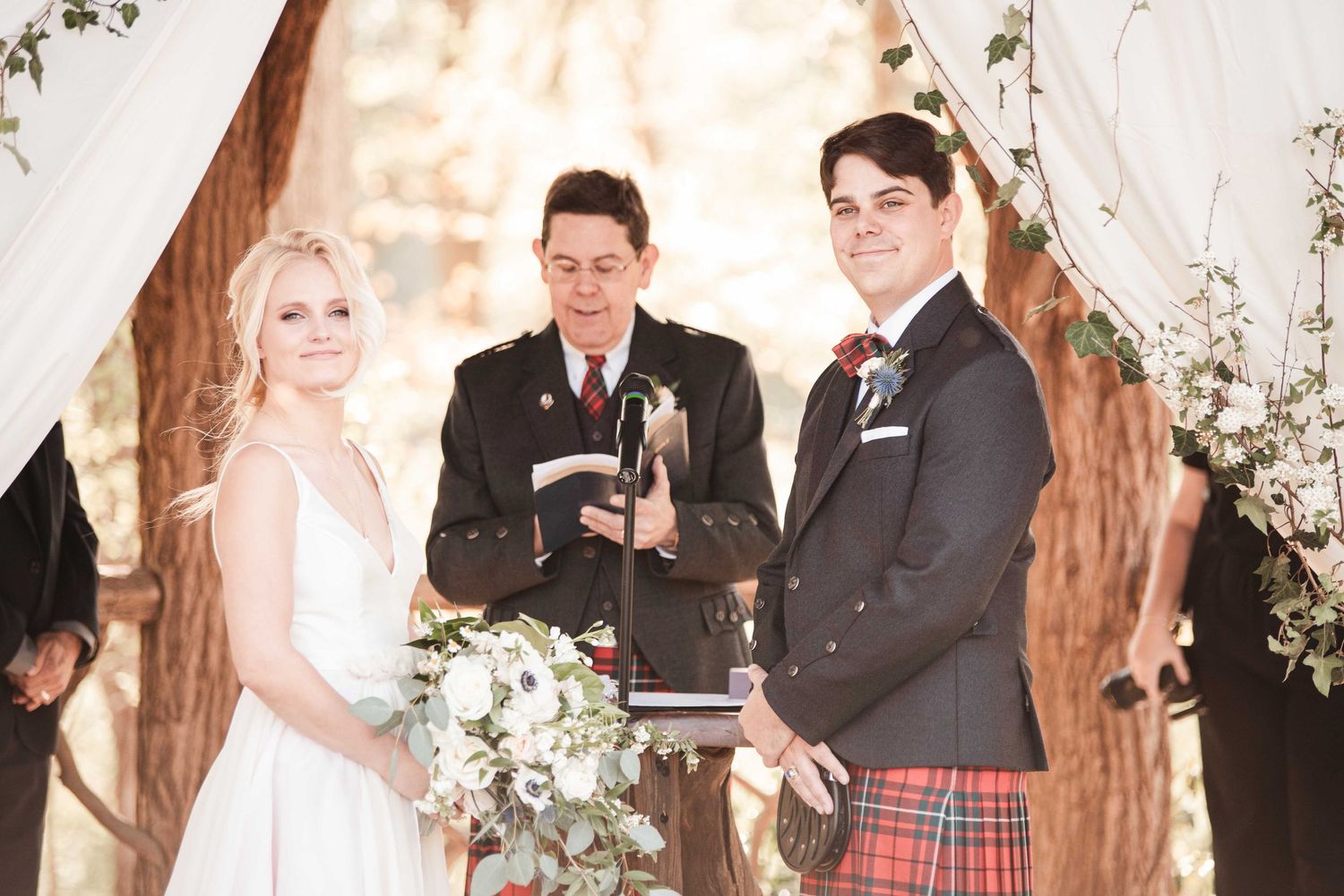 Bride and groom smiling at outdoor wedding ceremony with officiant, groom wearing red tartan kilt and boutonniere.