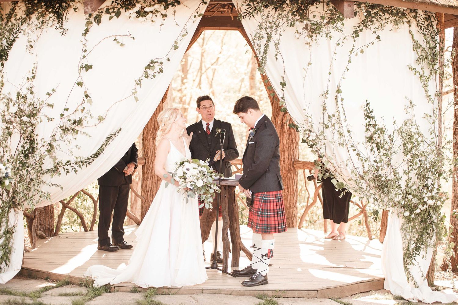 Bride and groom exchange vows under a floral arch with white draping and greenery at an outdoor wedding ceremony.