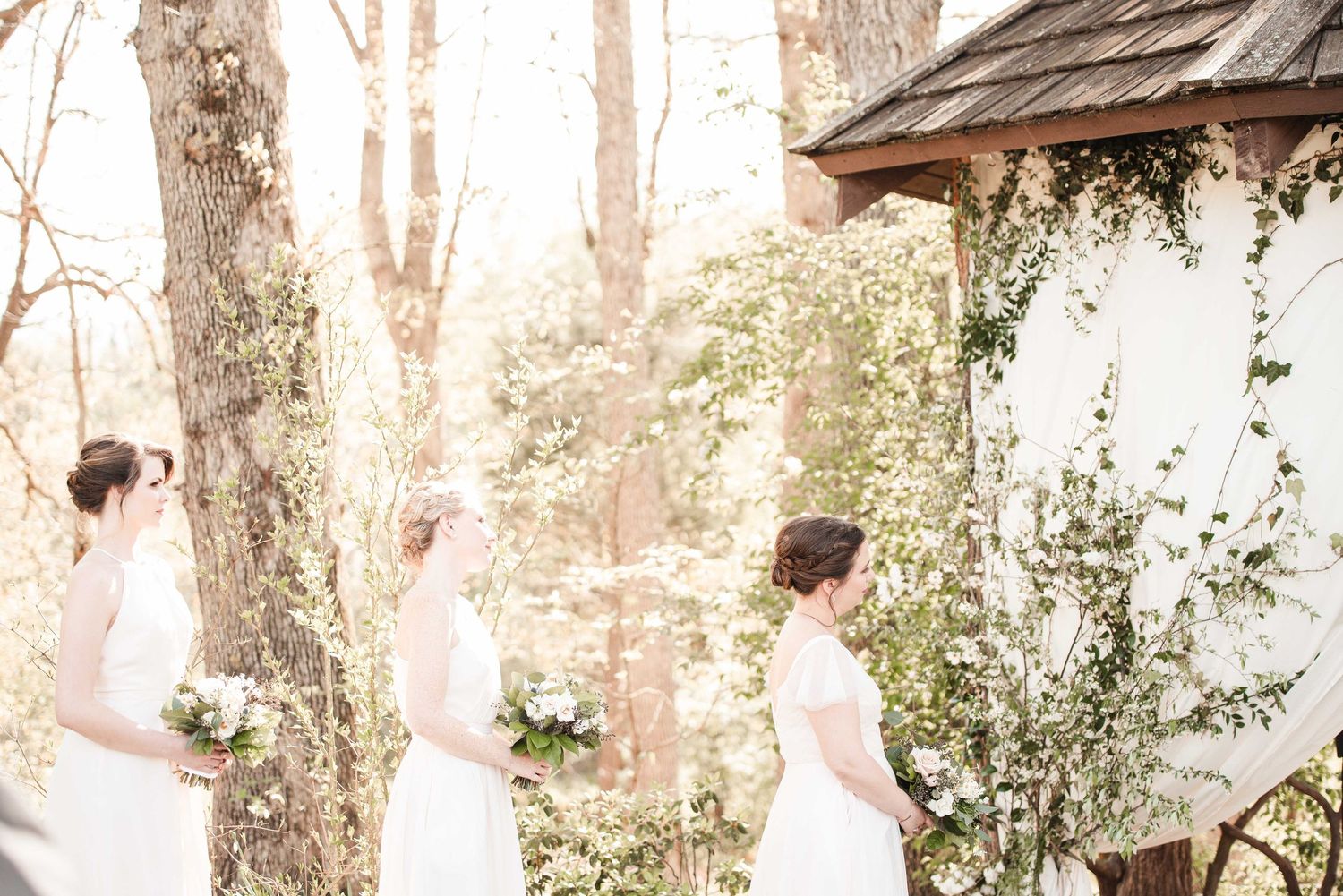 Three bridesmaids in white dresses holding bouquets stand among blooming trees at an outdoor spring wedding ceremony.