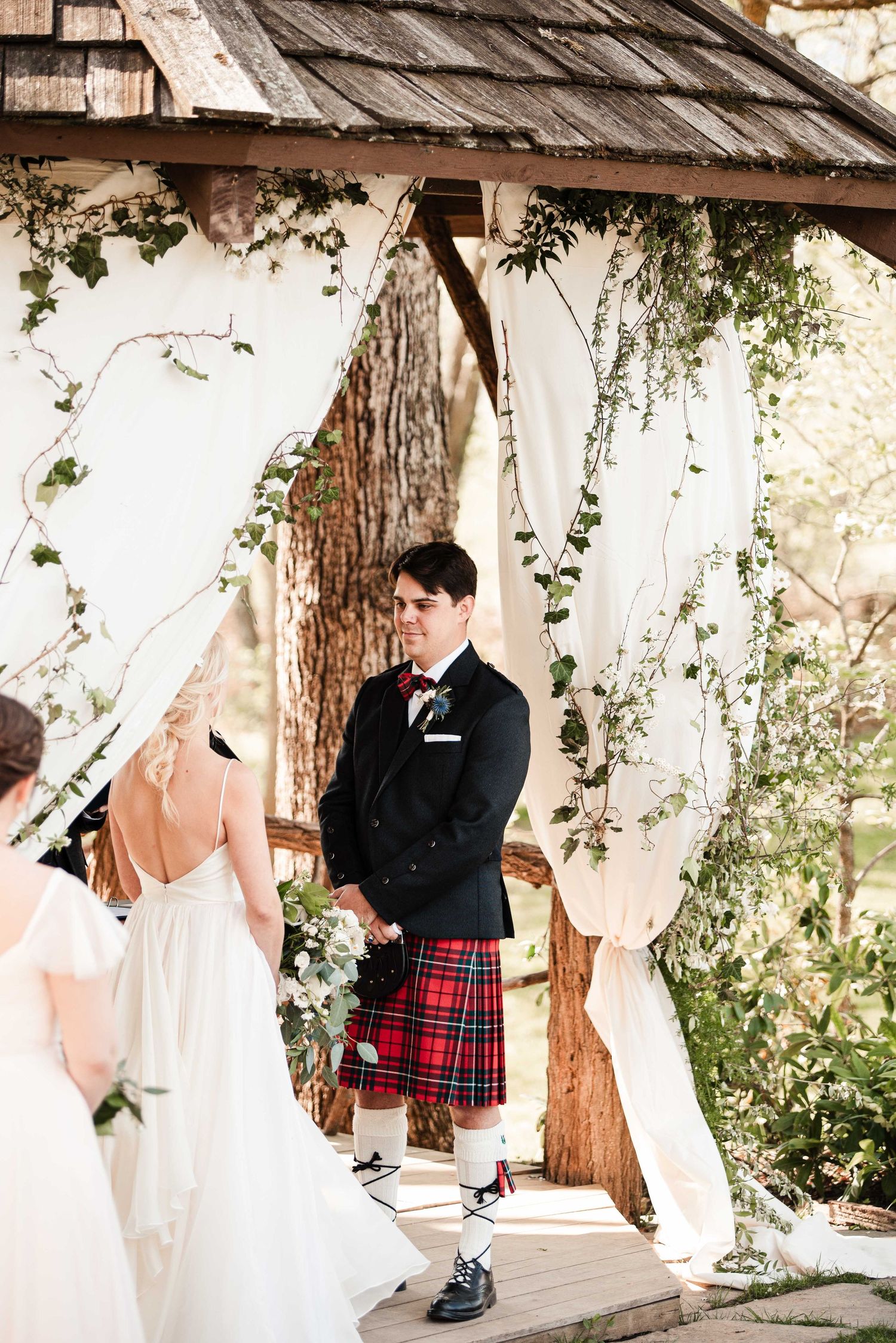 Groom in black jacket and red tartan kilt stands at outdoor wedding altar adorned with white draping and greenery.