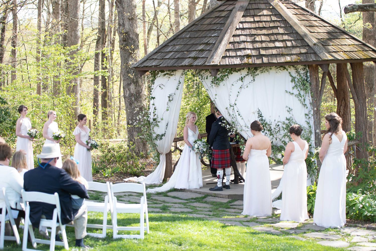 Outdoor wedding ceremony under a gazebo with bridesmaids in white dresses, surrounded by lush green trees.