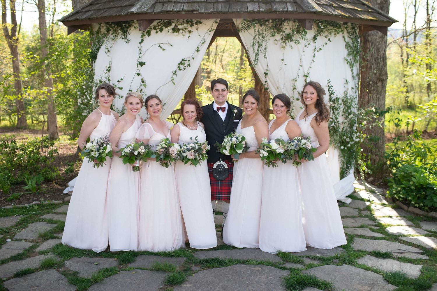 Groom in kilt with six bridesmaids in blush gowns holding bouquets under floral gazebo at outdoor wedding.