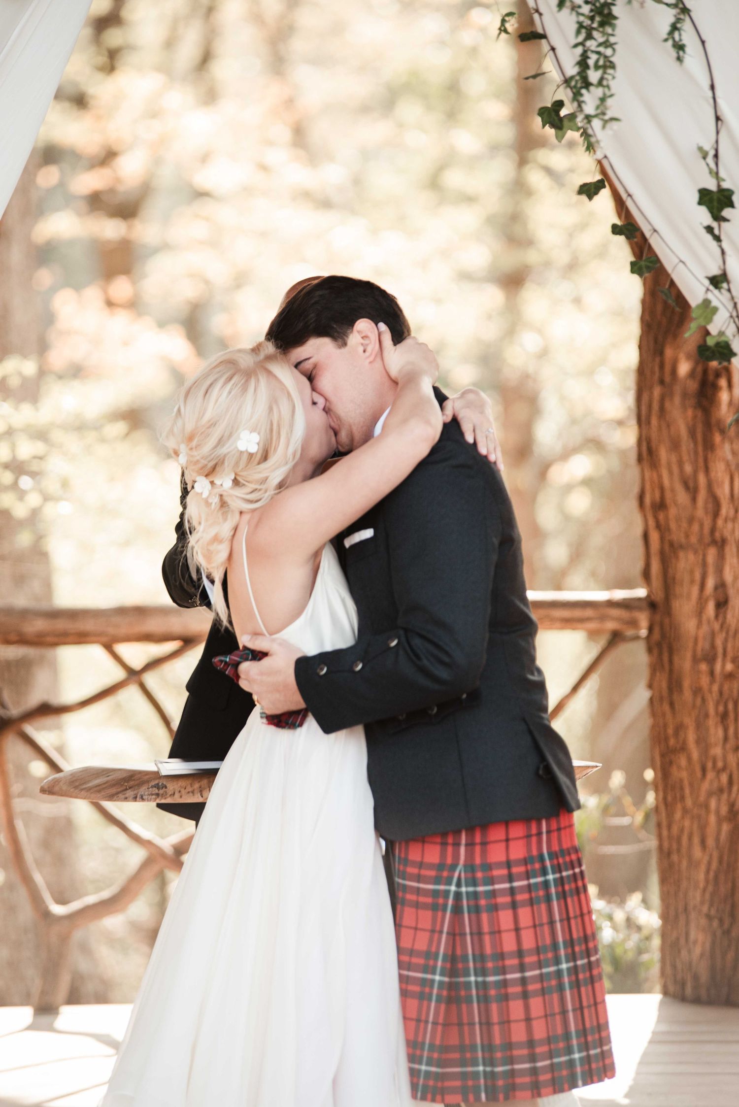 Bride and groom share first kiss at outdoor wedding ceremony, groom wearing traditional red tartan kilt and dark jacket.