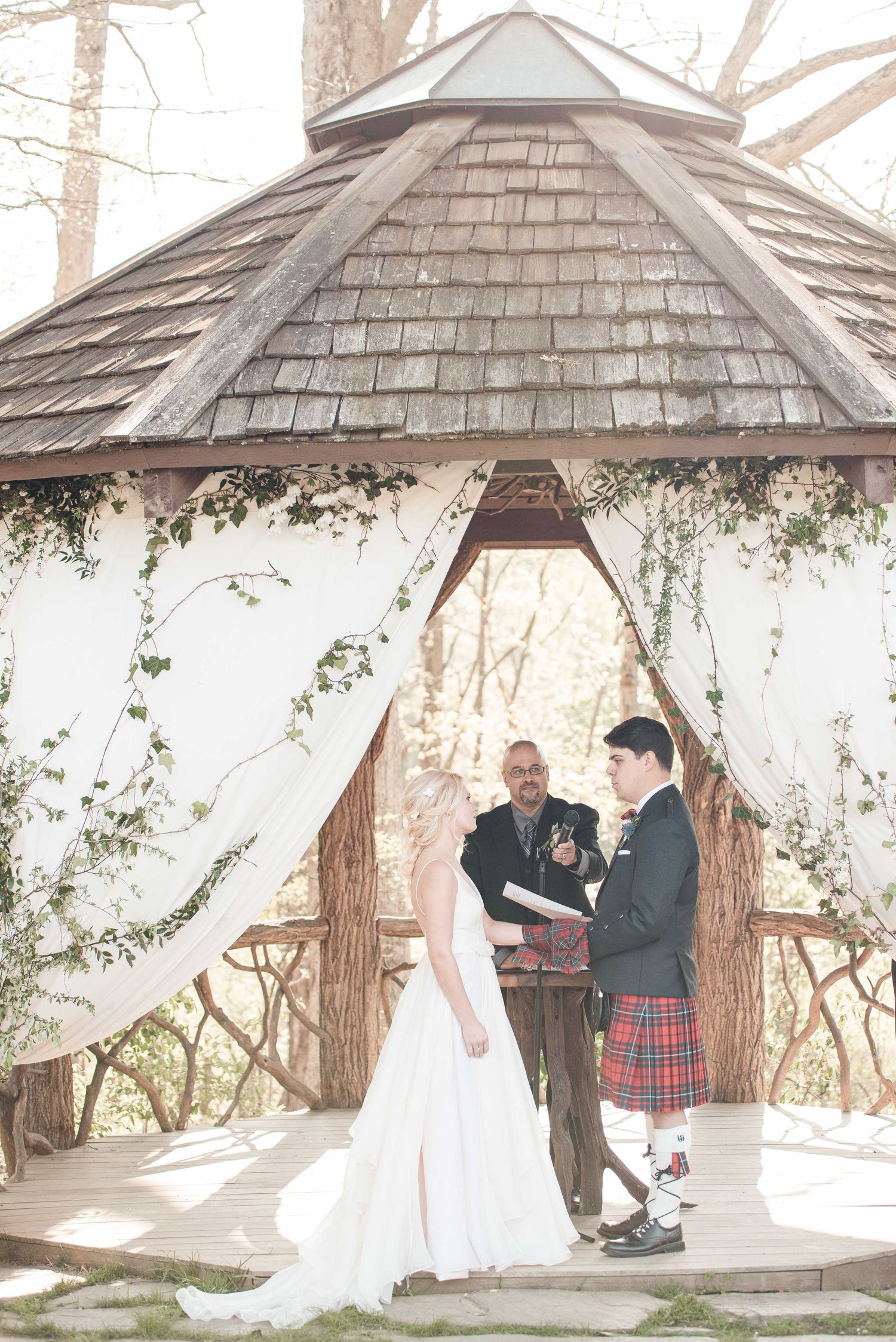 Bride and groom exchange vows under a decorated gazebo with white draping and ivy during an outdoor wedding ceremony.