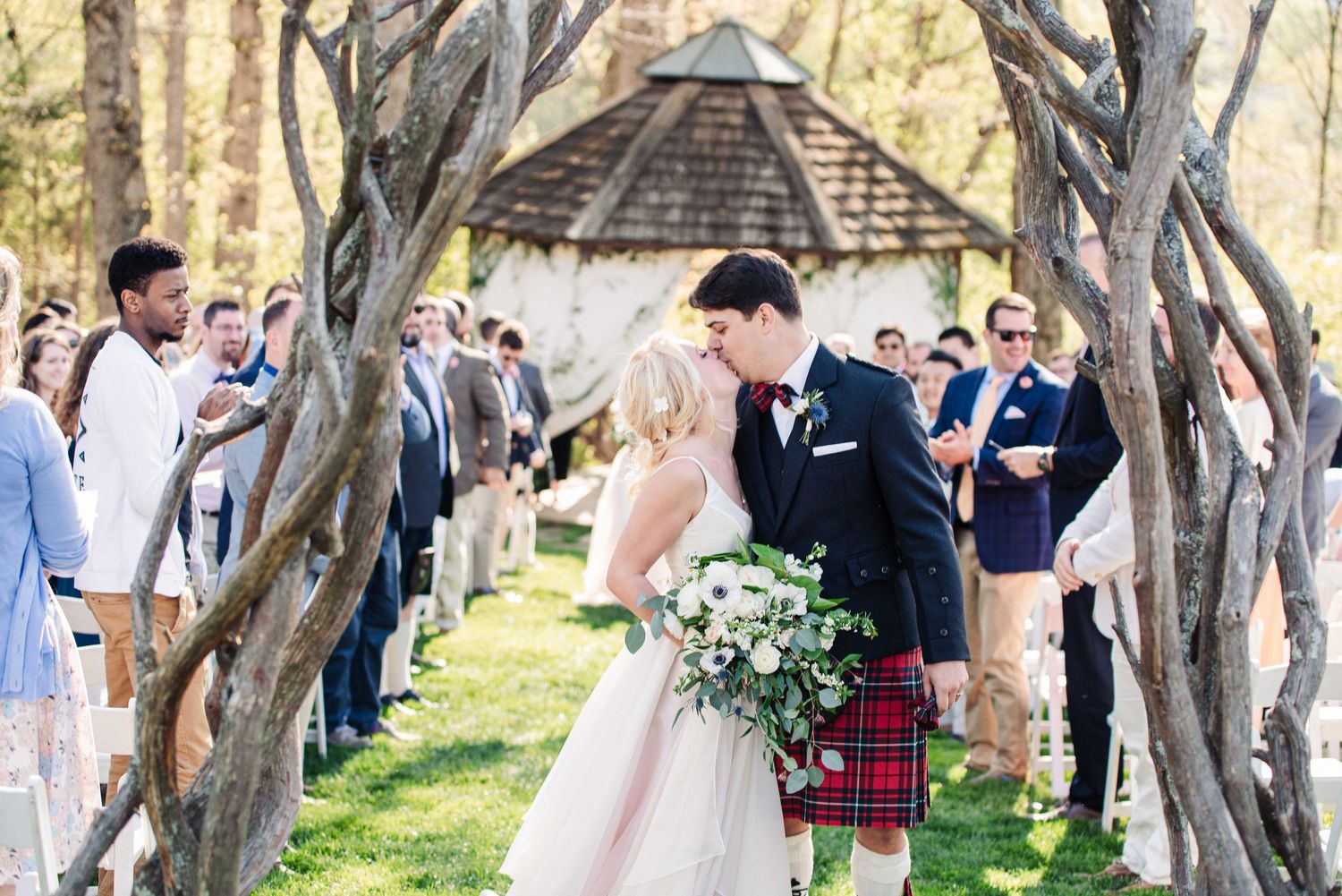 Bride and groom share a kiss at outdoor wedding ceremony, groom wearing red tartan kilt, surrounded by rustic tree branch arch.
