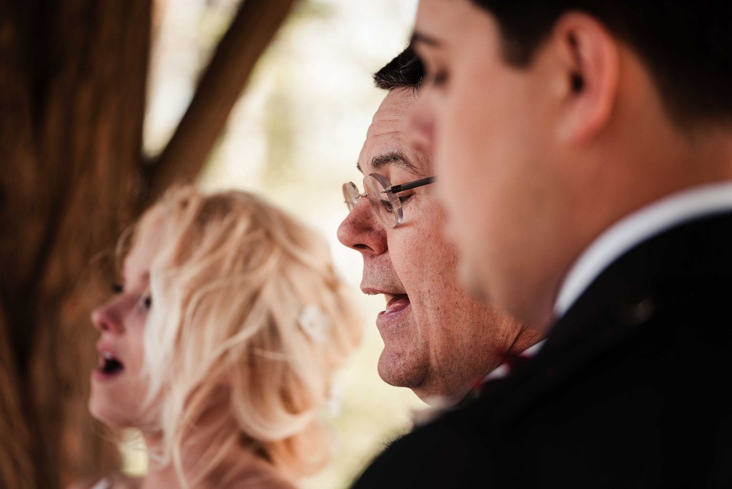 A man wearing glasses smiles warmly at a wedding ceremony, with a blonde woman visible in the background.