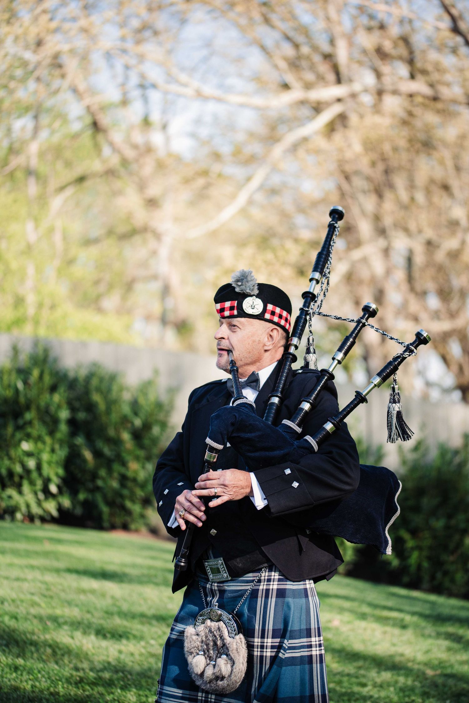 A Scottish bagpiper in traditional tartan kilt and beret performs outdoors in a lush green garden setting.