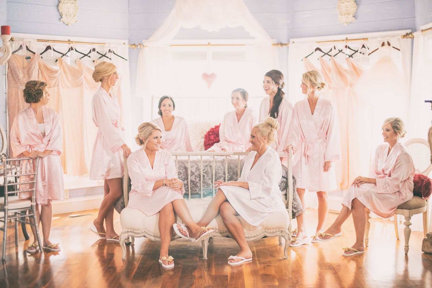 Bride and bridesmaids in matching pink robes relax together in a bright, elegant bridal suite.