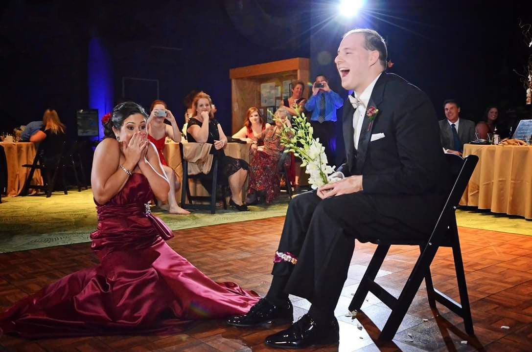 A woman in a red gown sits on the floor laughing with a man in a tuxedo holding flowers at a formal event.