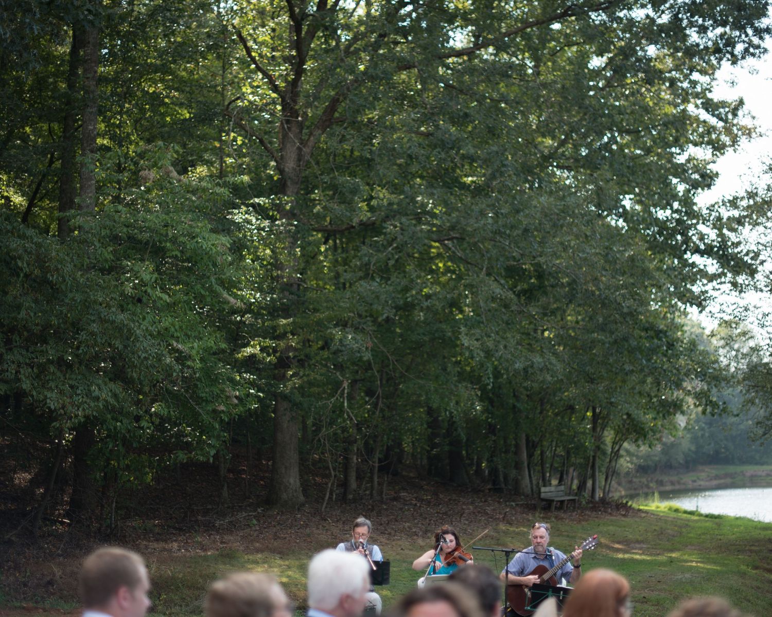 Outdoor wedding ceremony in a wooded setting near a river with guests seated among tall trees.