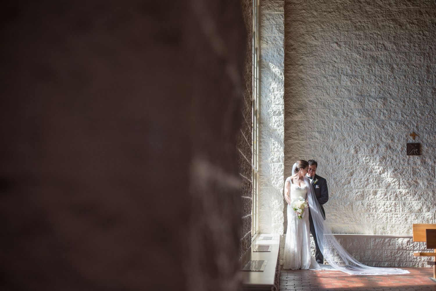 A bride and groom share a romantic kiss framed by a stone wall and streaming sunlight on their wedding day.