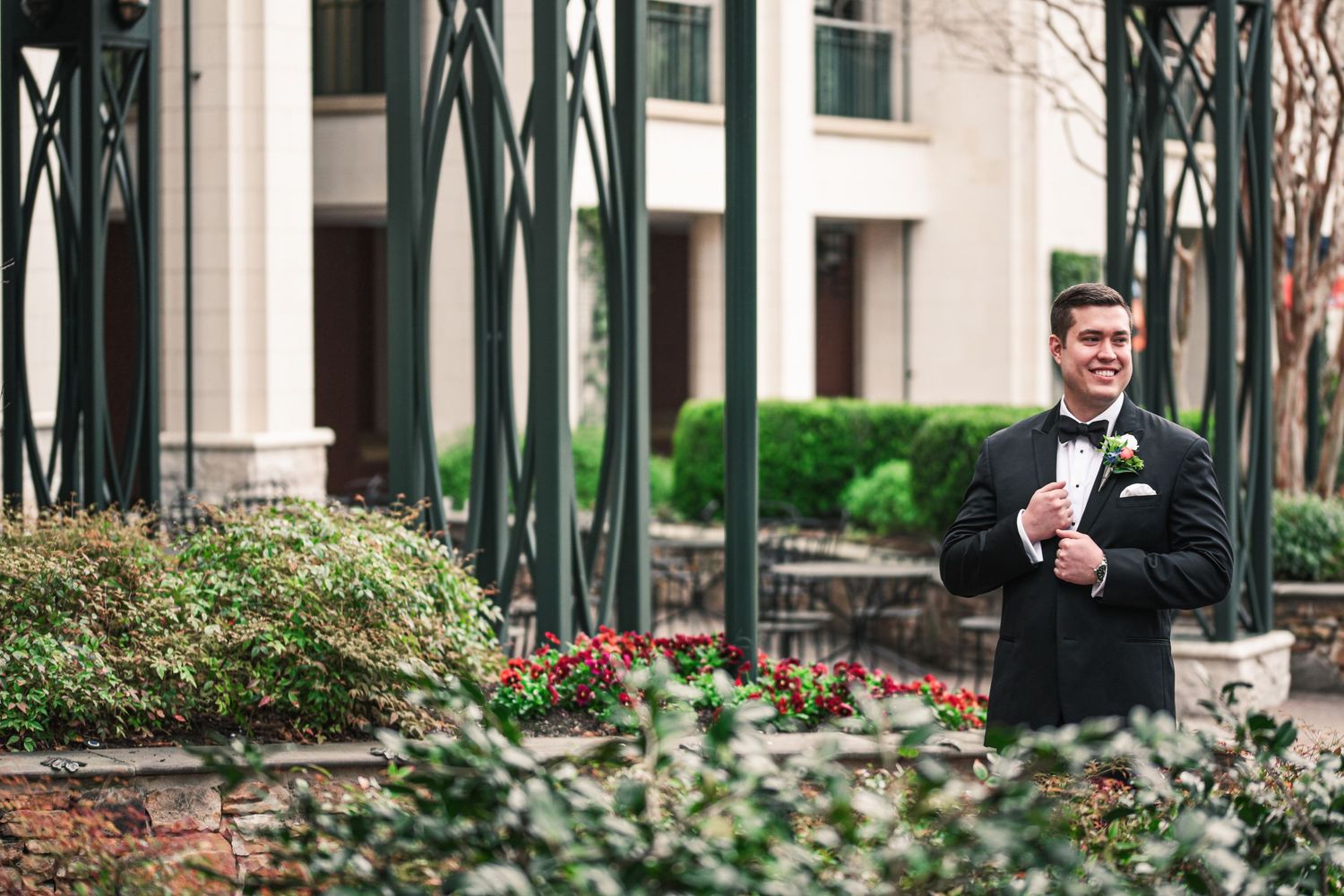 Dapper groom in black tuxedo with boutonniere smiling outdoors at garden wedding venue.