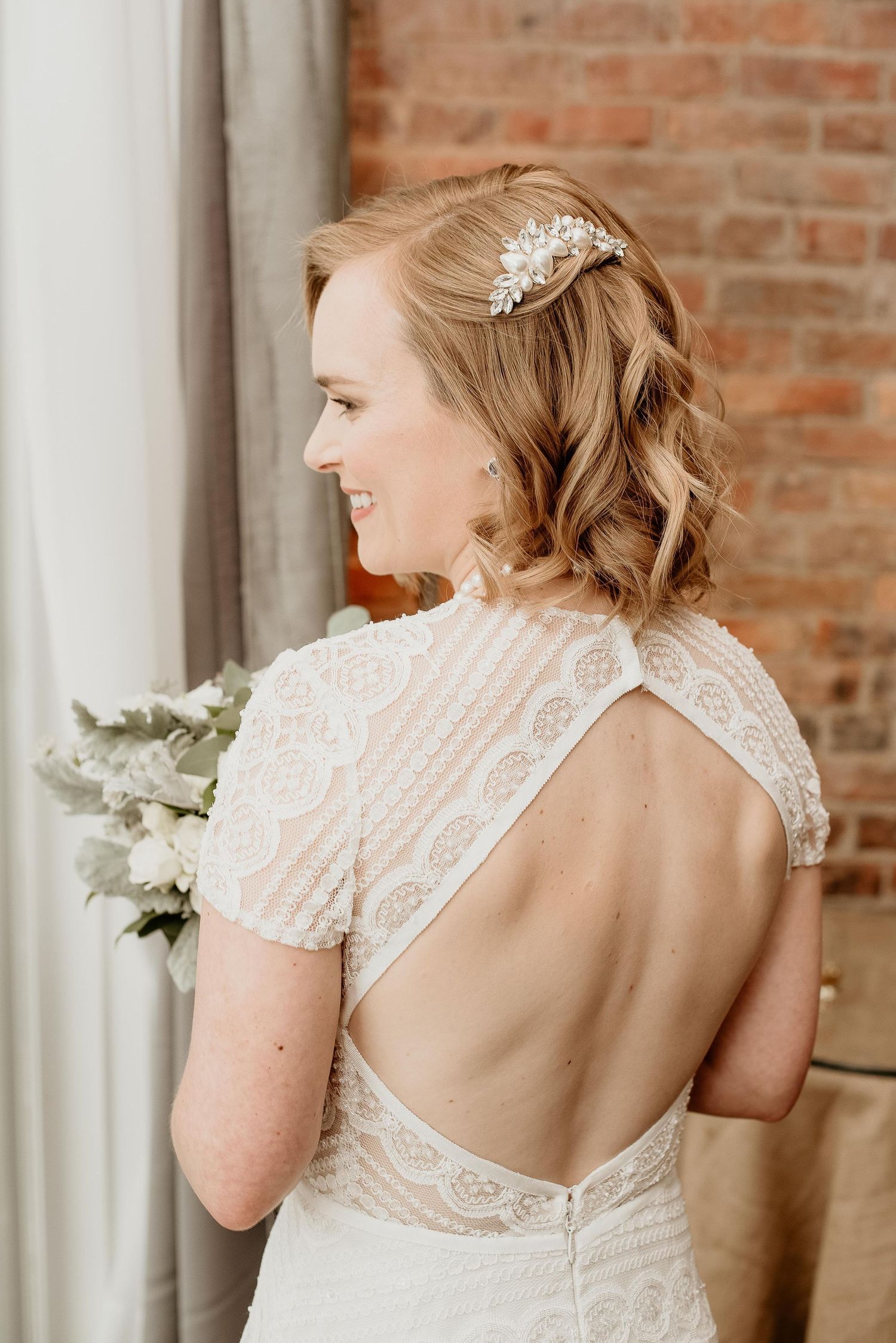 Bride in open-back lace wedding gown with floral hair accessory, smiling over shoulder near brick wall.