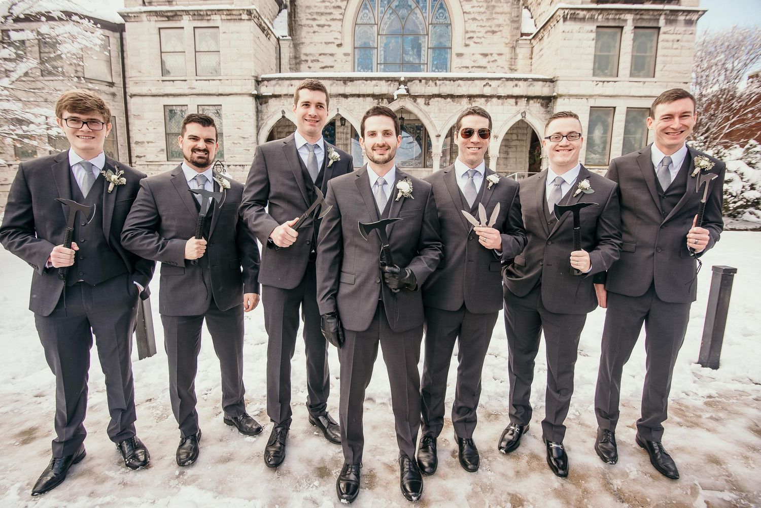 Seven groomsmen in dark suits pose outdoors in snow near a stone church building on a winter wedding day.