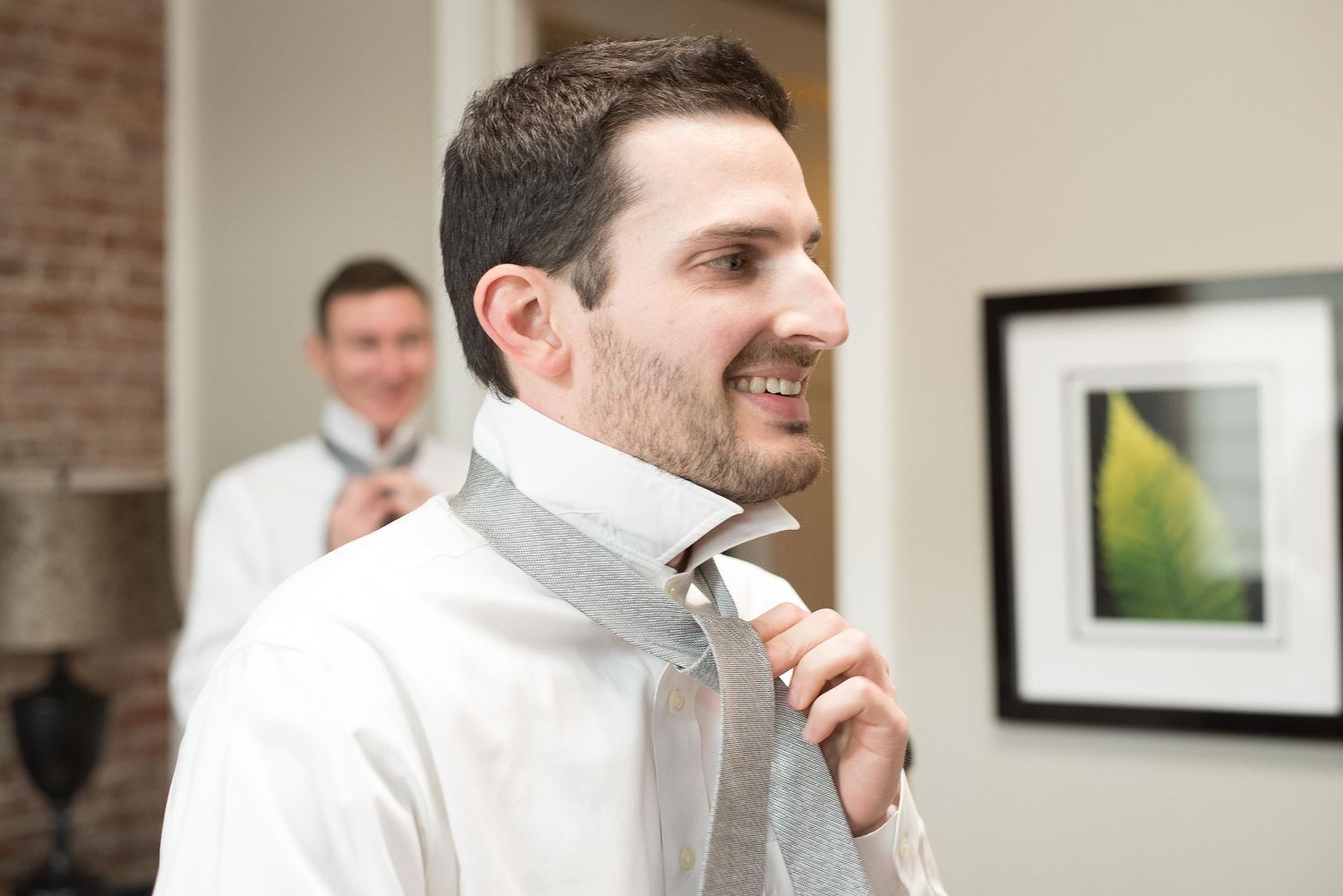 Smiling groom adjusting his gray tie while getting ready for his wedding ceremony.