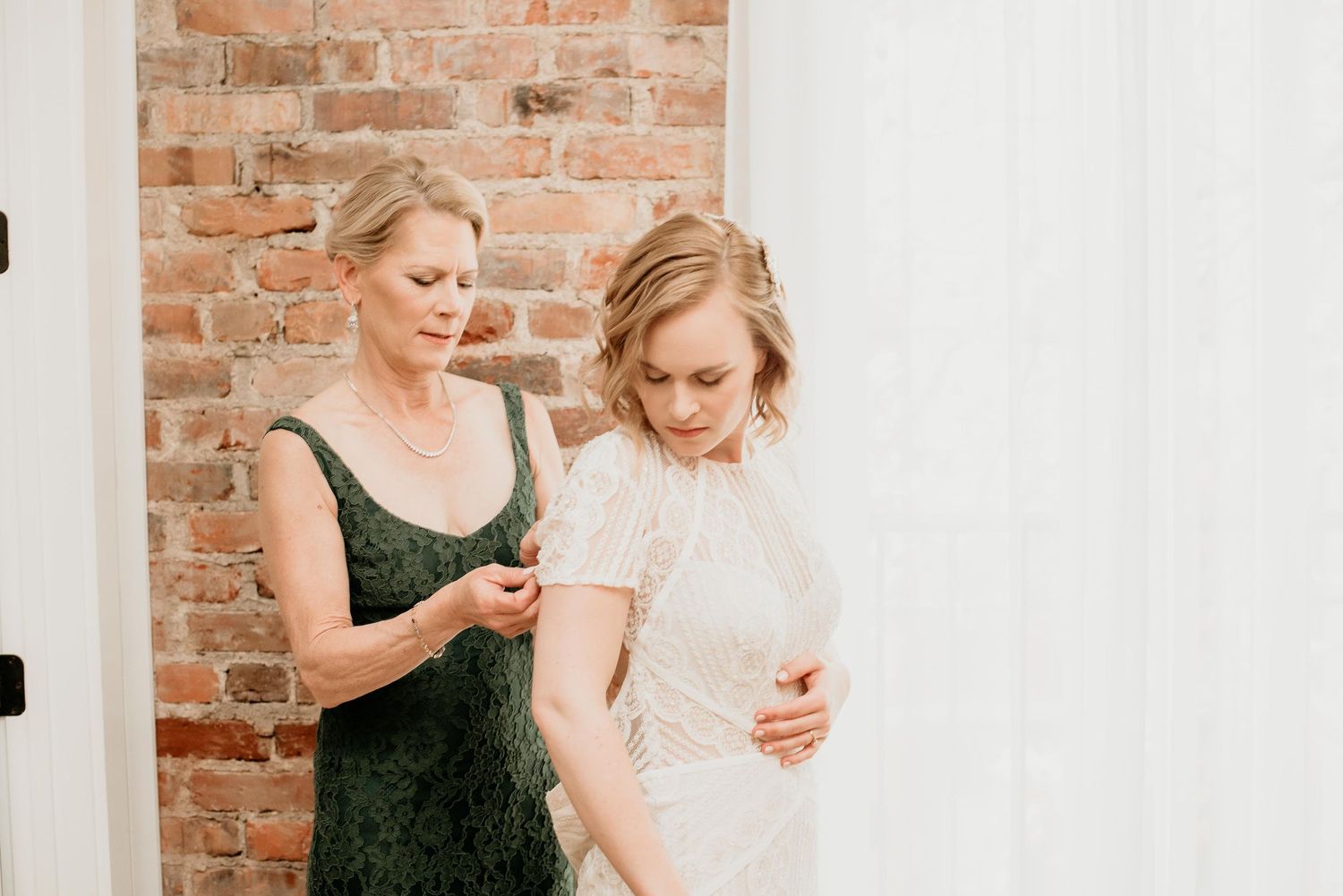 Mother helping bride button her lace wedding dress in a bright room with exposed brick wall backdrop.