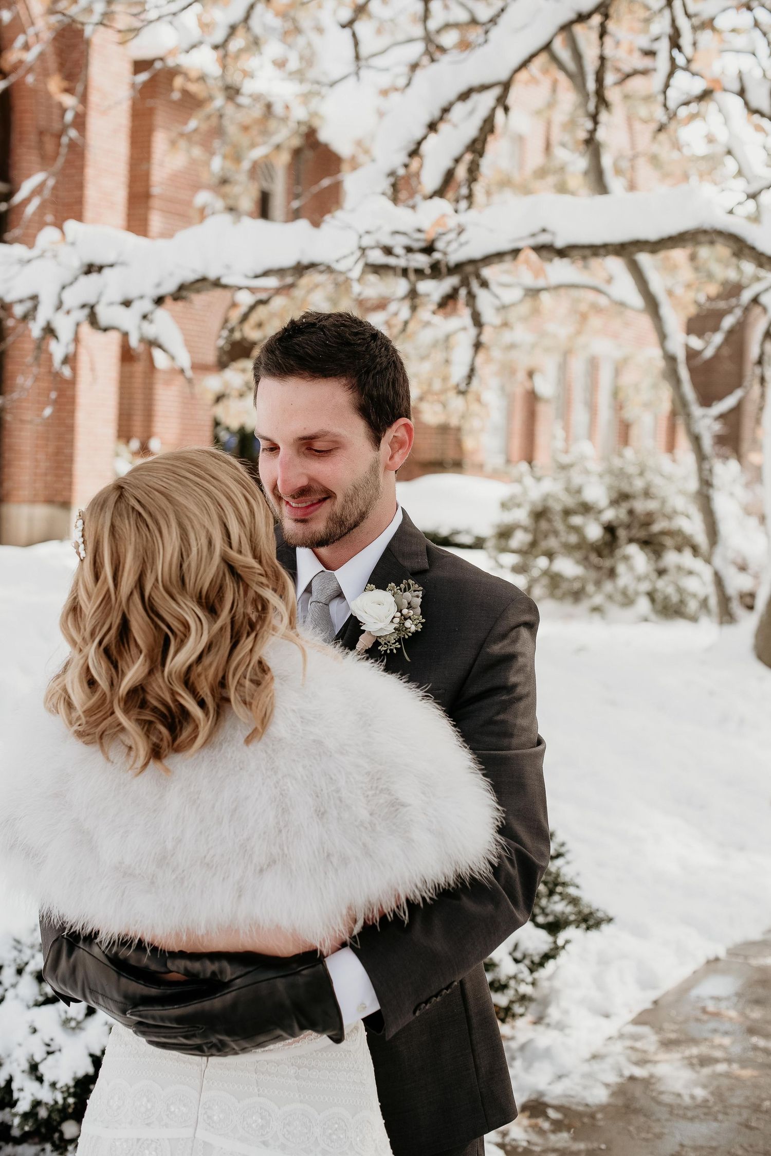 Bride in white fur stole embraced by groom in gray suit during a romantic winter wedding outdoors in the snow.