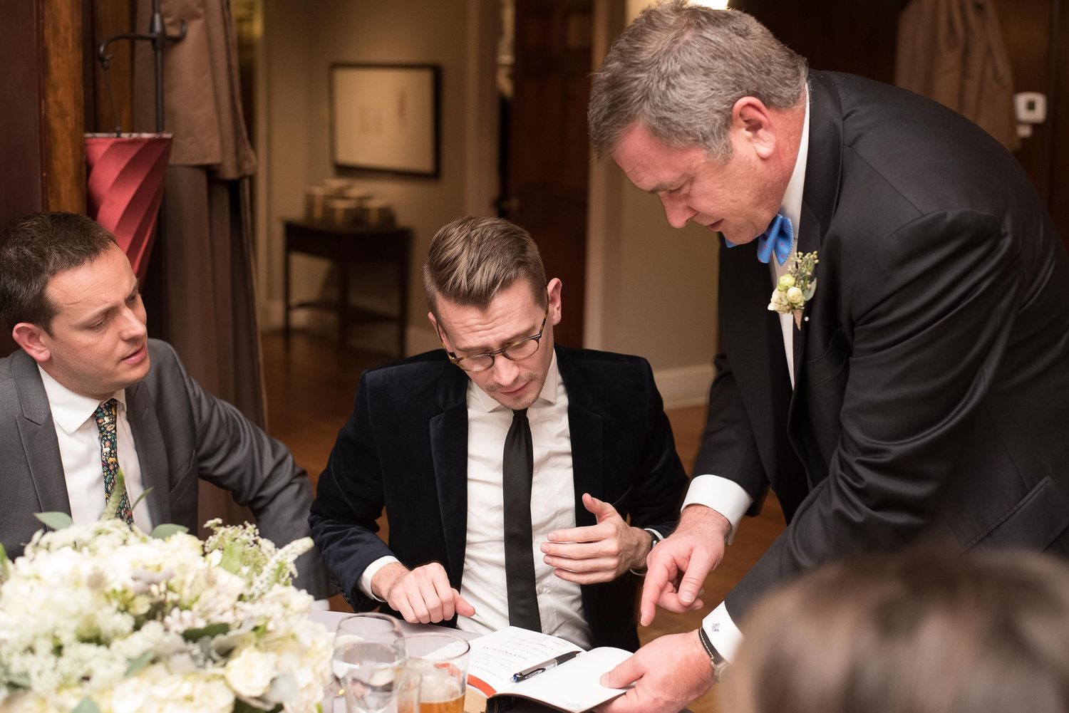 Men in formal attire reviewing documents at an elegant wedding reception table with white floral arrangements.