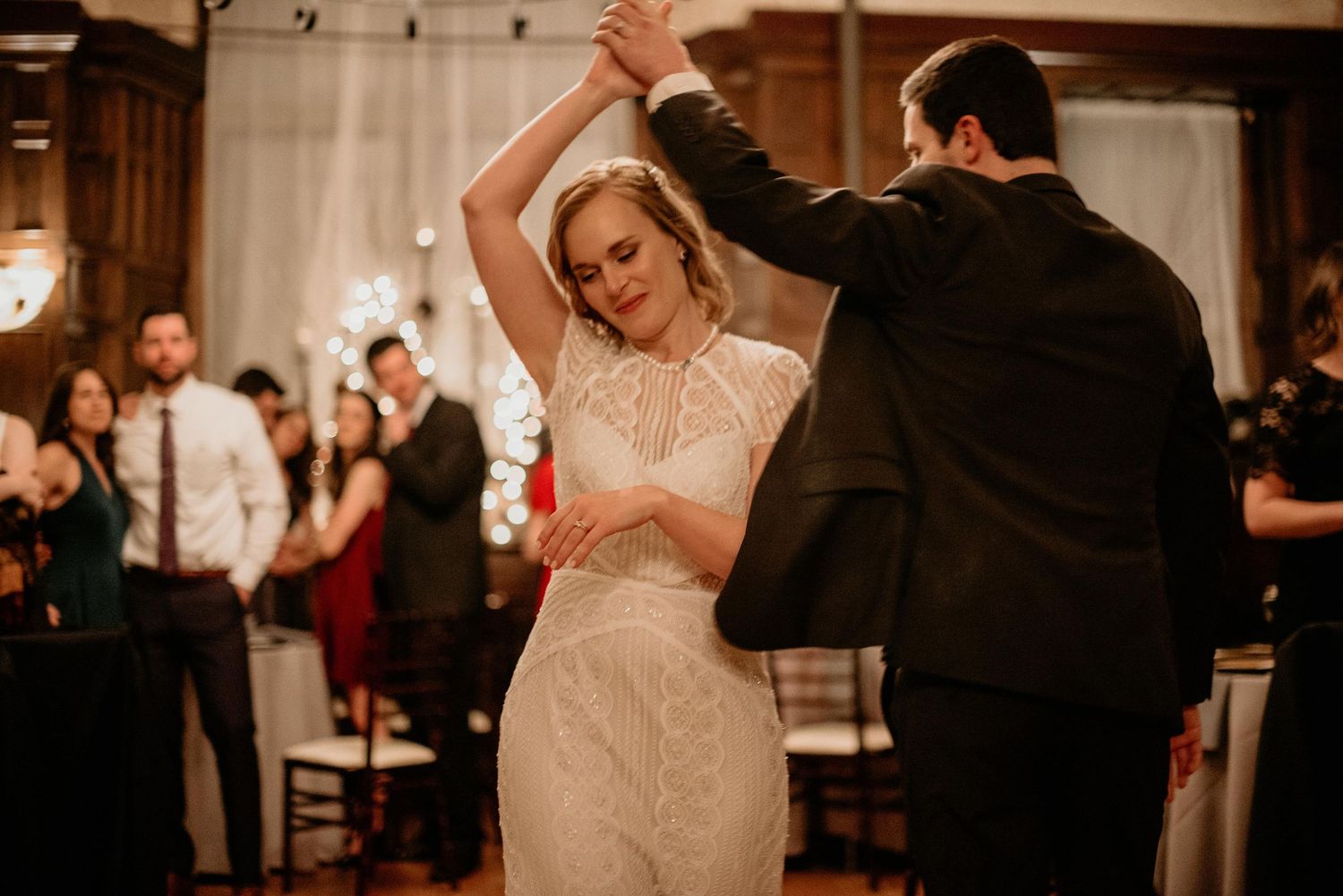 Bride in lace gown and groom in black tuxedo share first dance at elegant wedding reception.